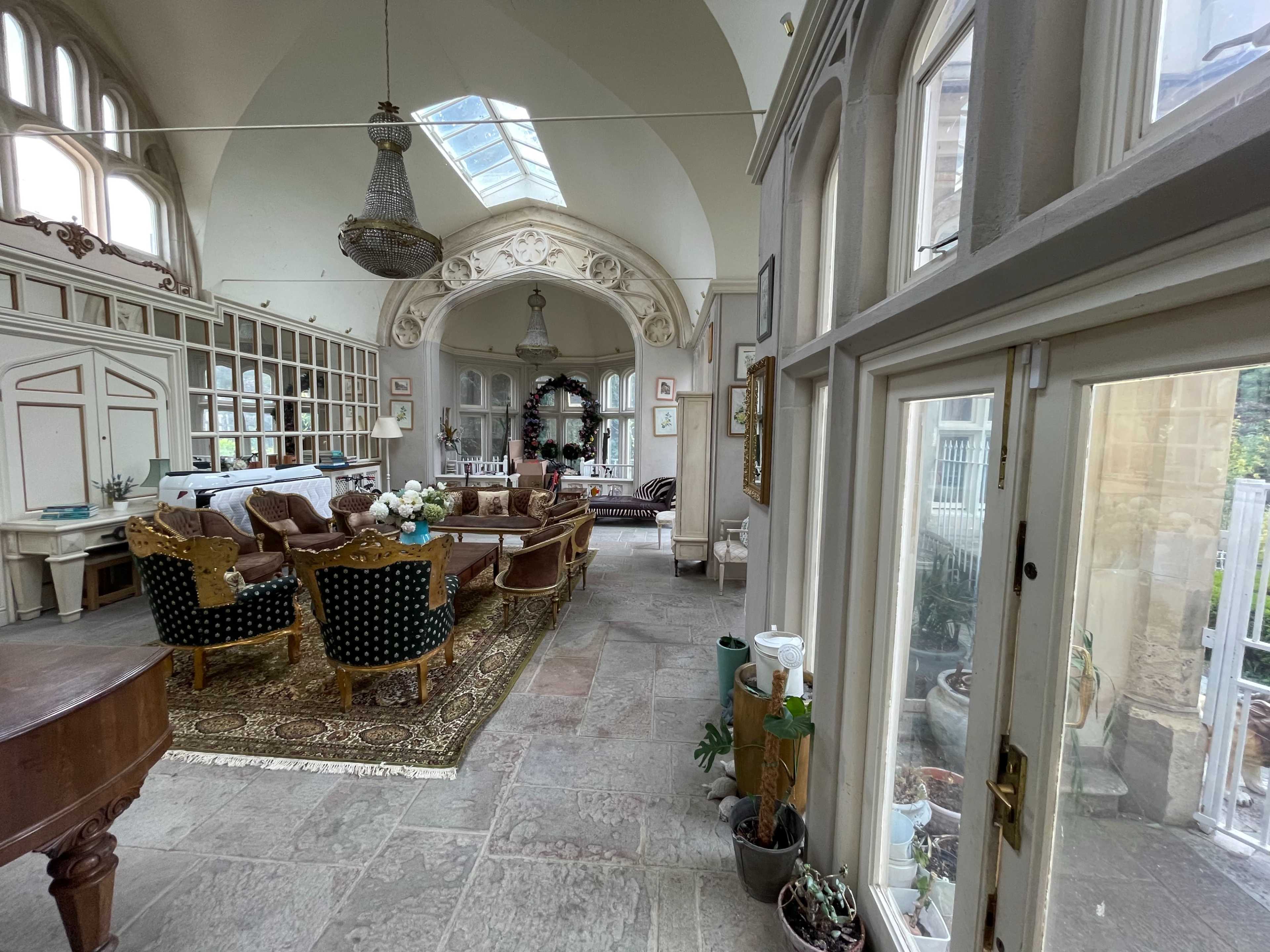 The image shows a spacious, elegantly decorated living room with a mix of vintage and modern furnishings, large windows, and a view of an outdoor area.