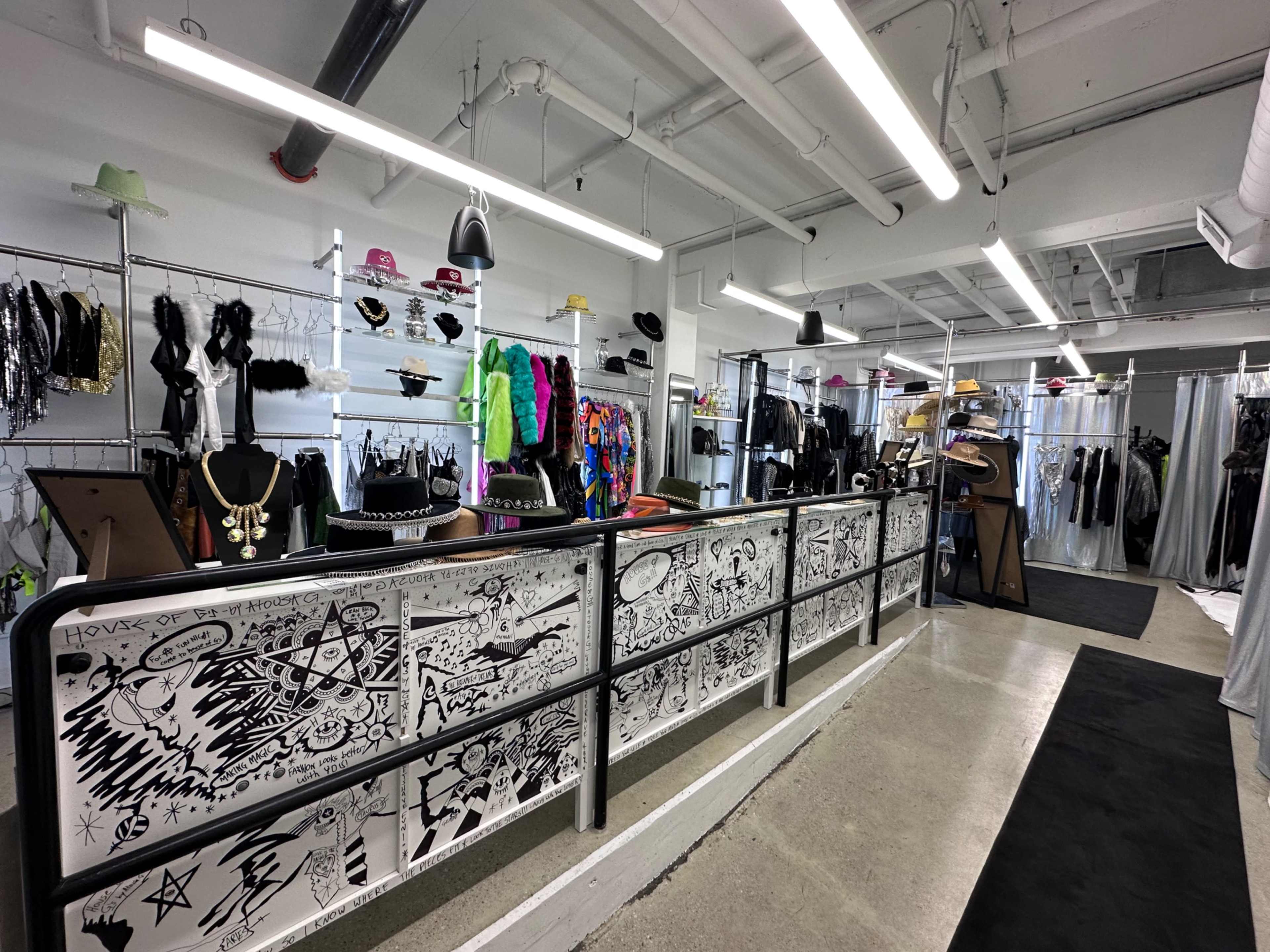The image shows a clothing store interior with a display of varied garments on racks, a checkout counter with artistic graffiti, and bright lighting.