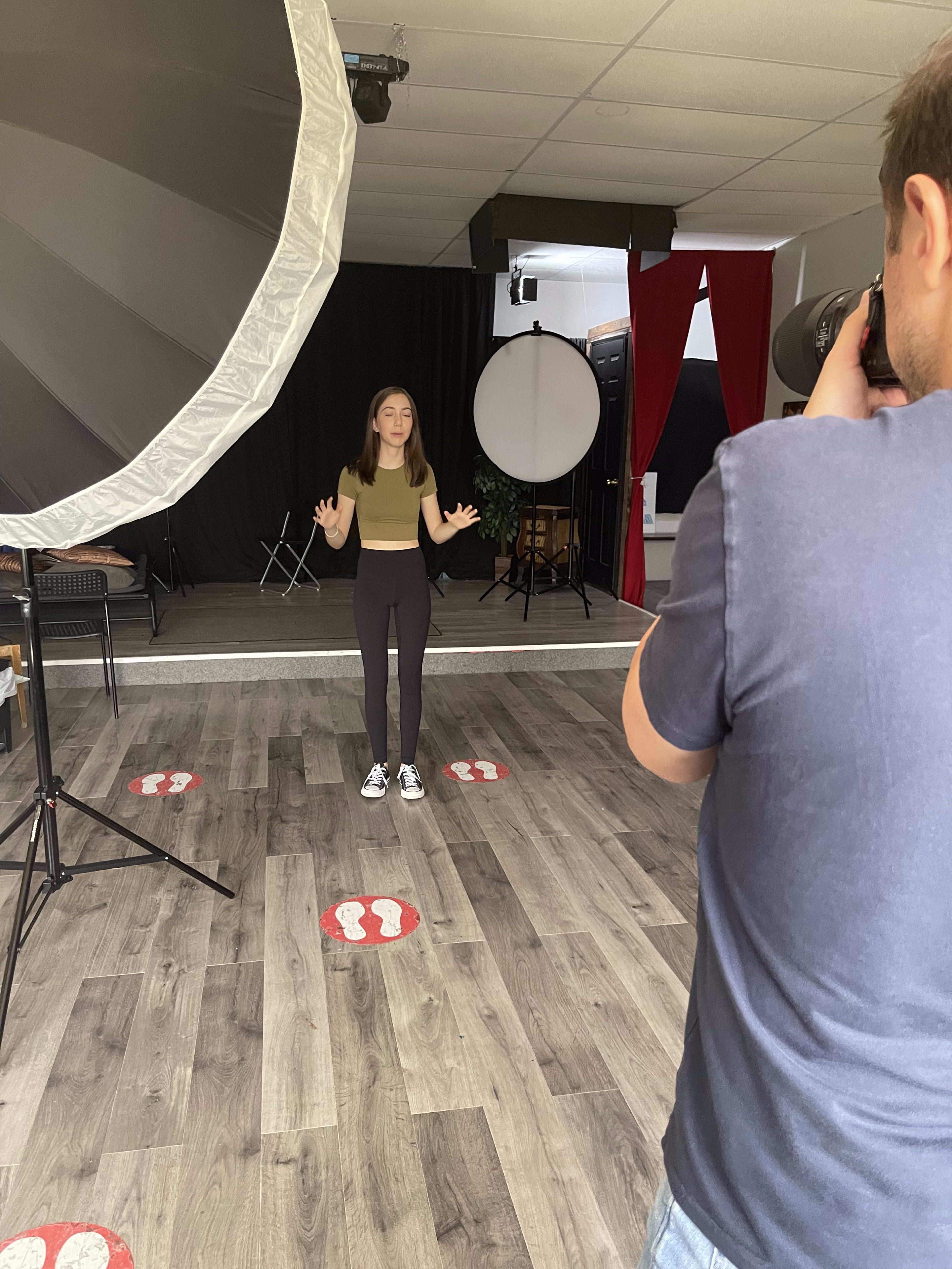 A young woman stands in a photography studio, while a person behind a camera prepares to take her picture.