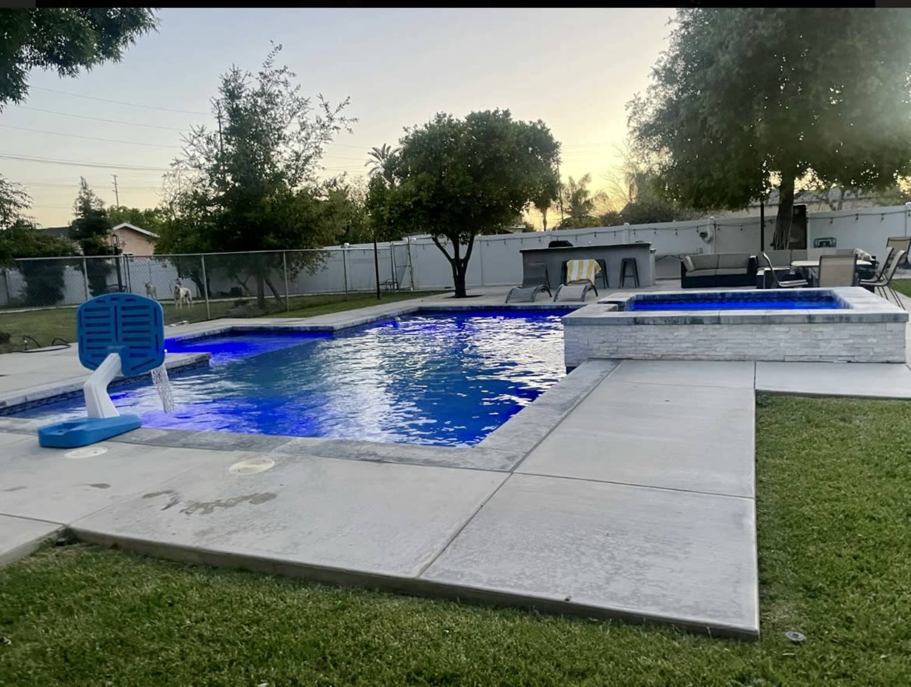 A backyard features a swimming pool with blue lighting and a hot tub beside it, surrounded by green grass and trees.