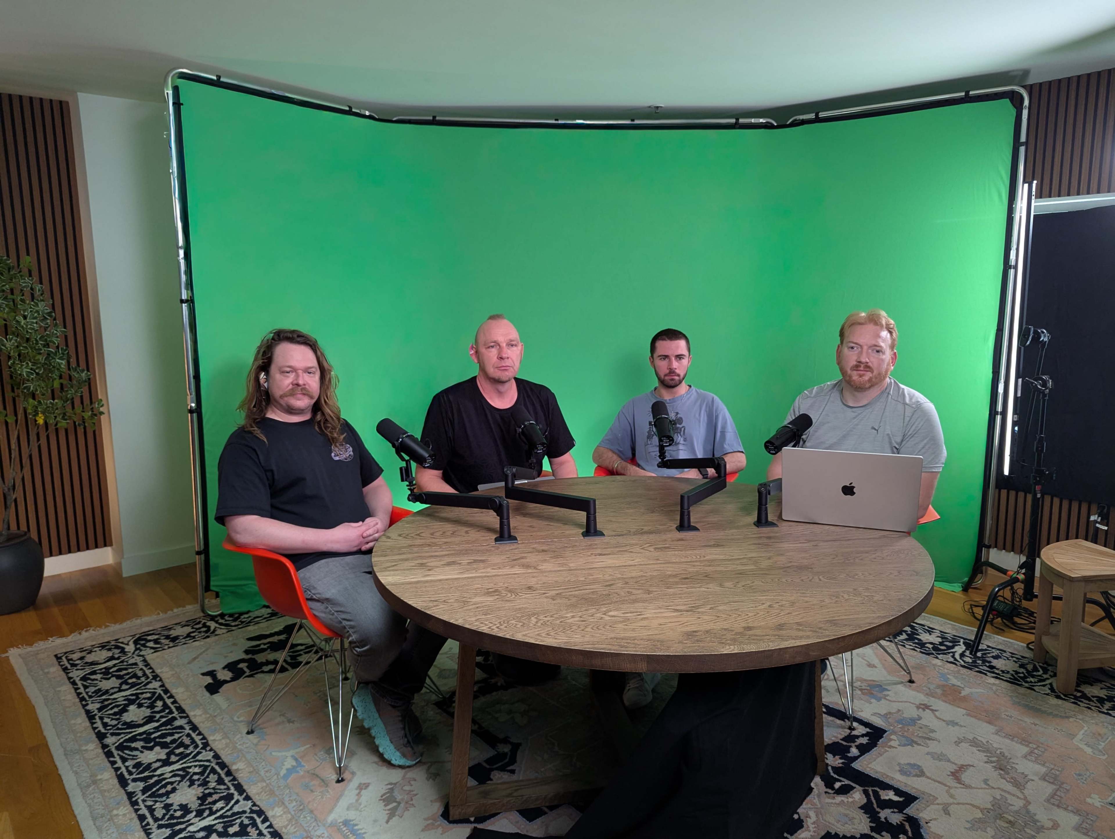 Four men are seated around a circular wooden table in front of a green screen, with microphones set up for a discussion.