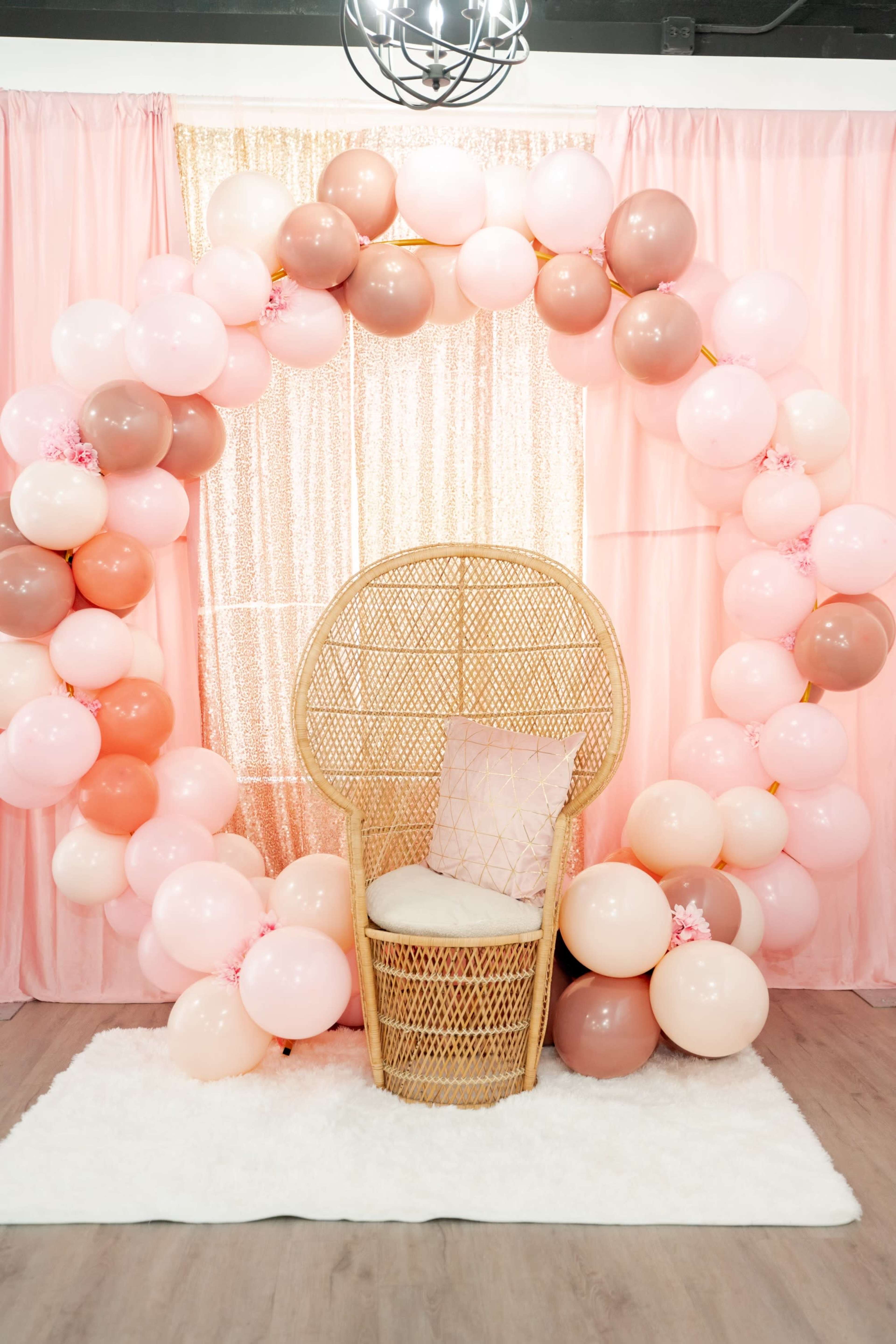 A rattan chair is placed in front of a large balloon arch, composed of pastel-colored balloons, with a pink backdrop and a plush white rug beneath.
