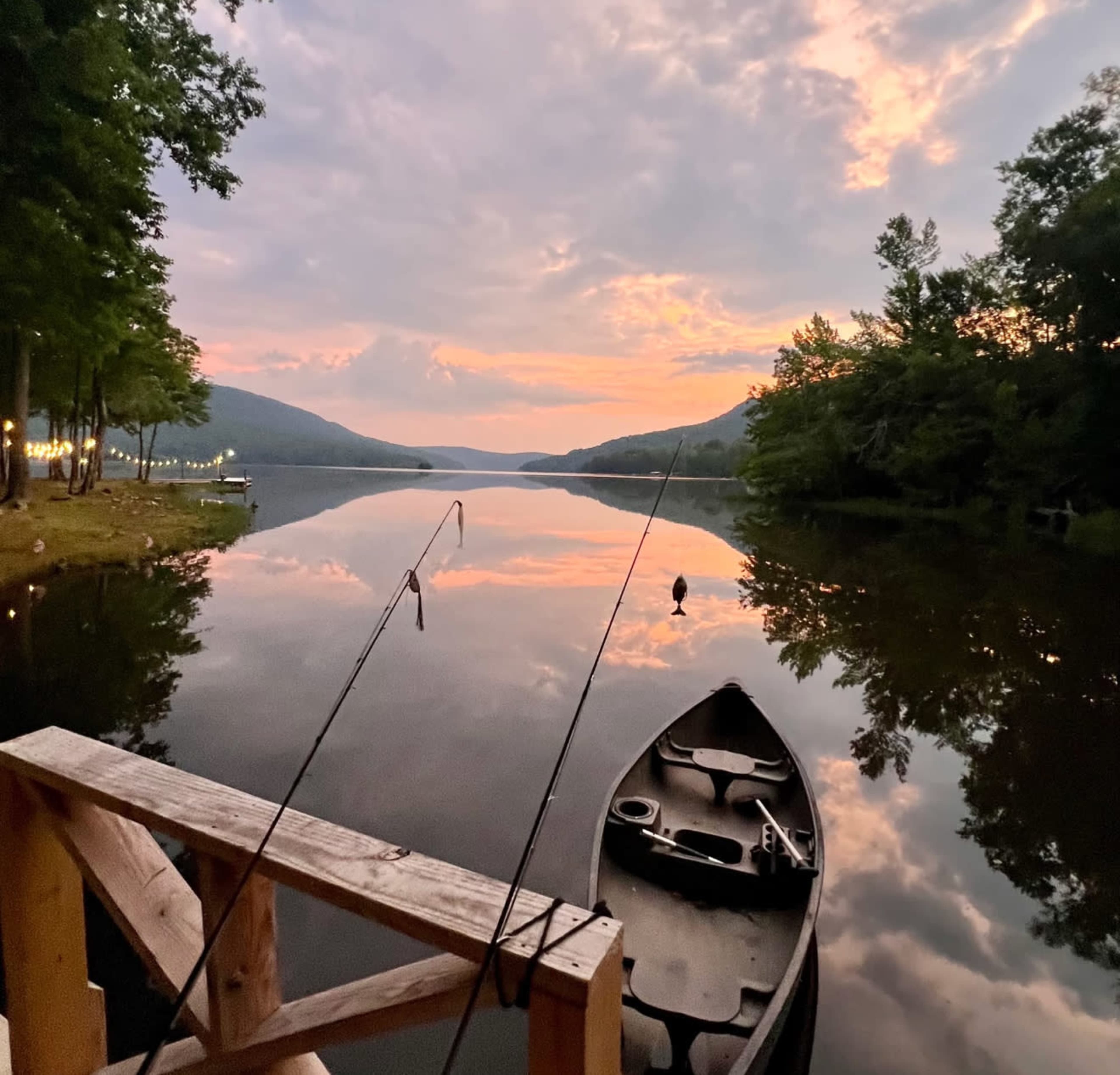 A calm lake reflects the colorful sky at dusk, with a fishing boat and rods visible on a wooden dock.