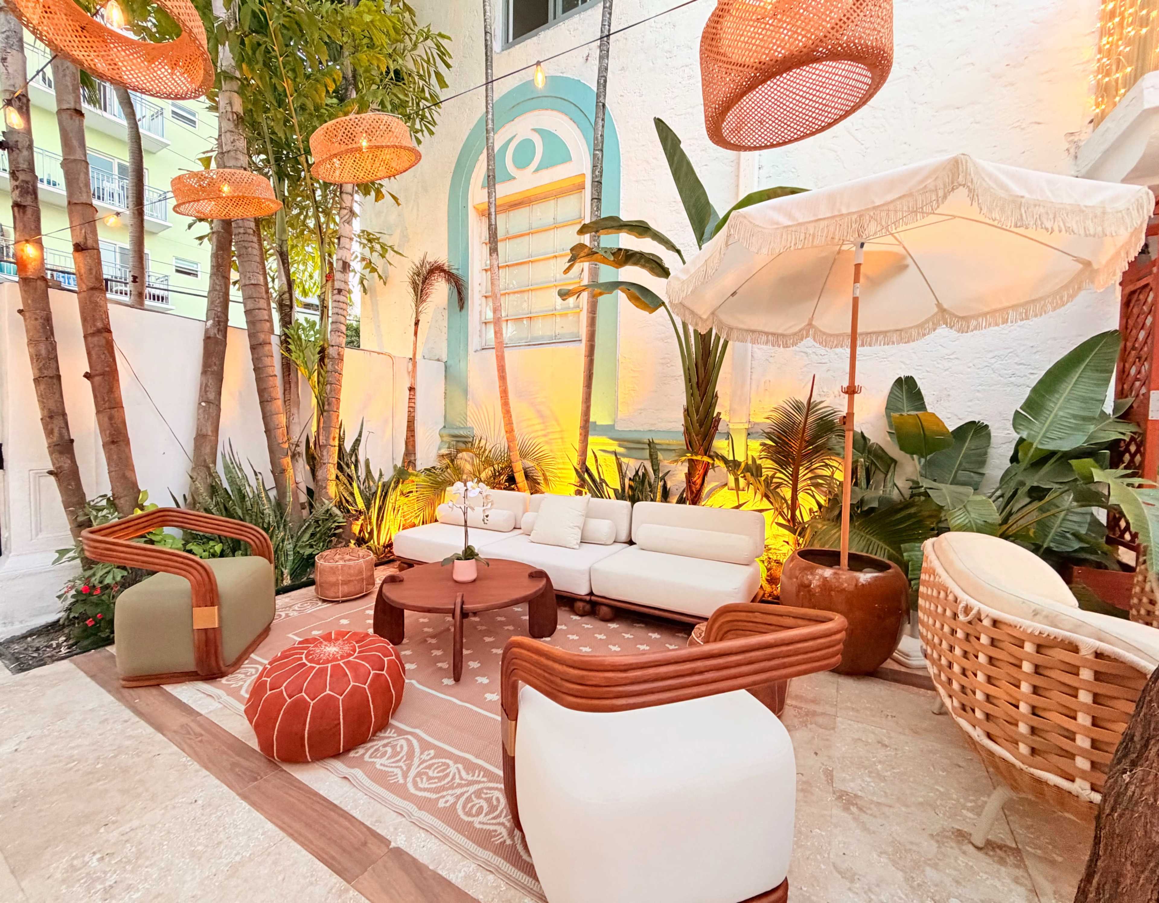 The image depicts a cozy outdoor lounge area with a white sofa, wooden furniture, potted plants, and decorative hanging light fixtures, surrounded by tropical foliage.