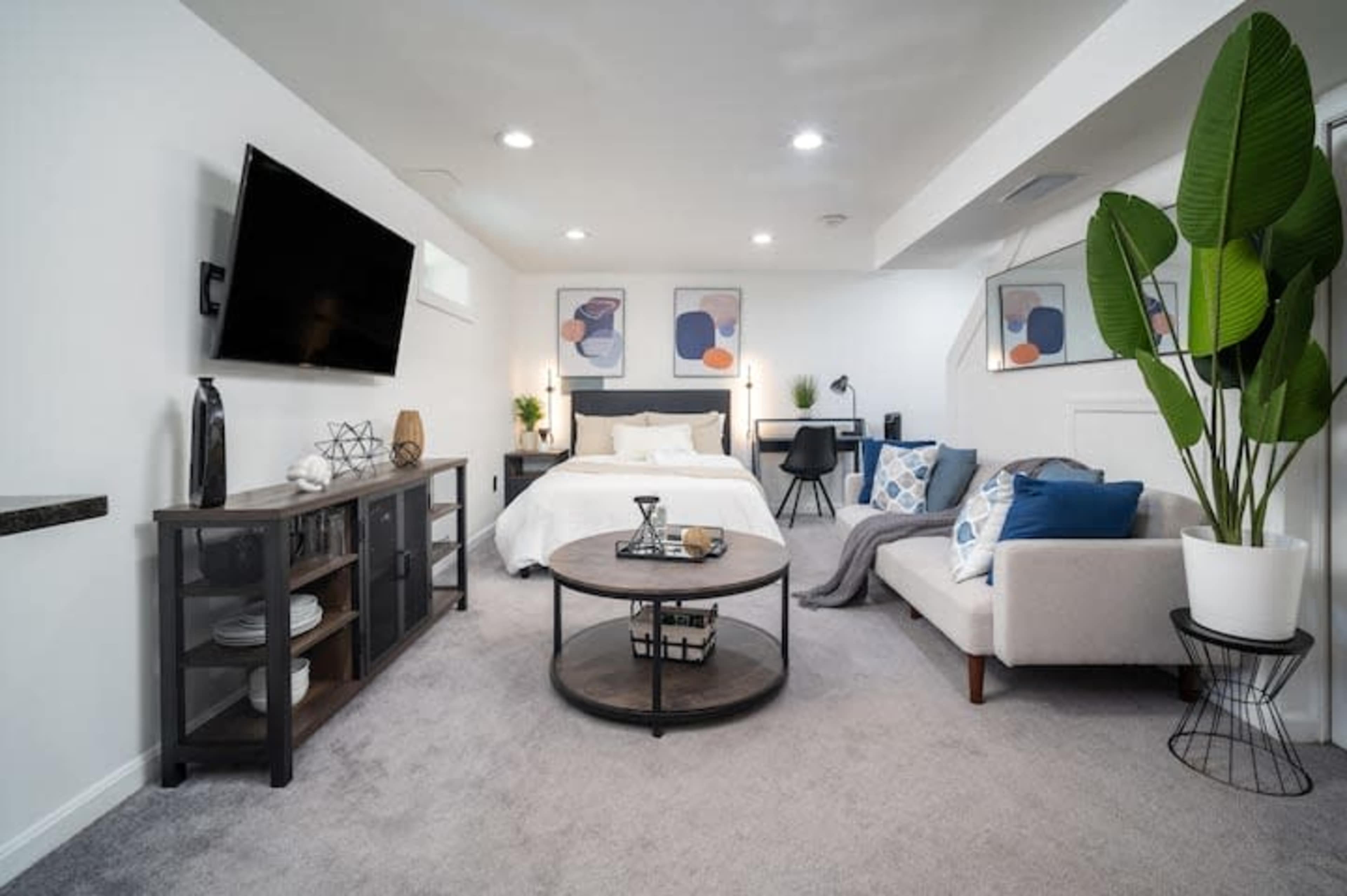 A modern basement bedroom features a bed, a sofa, a coffee table, and wall-mounted TV, surrounded by light-colored walls and decor.
