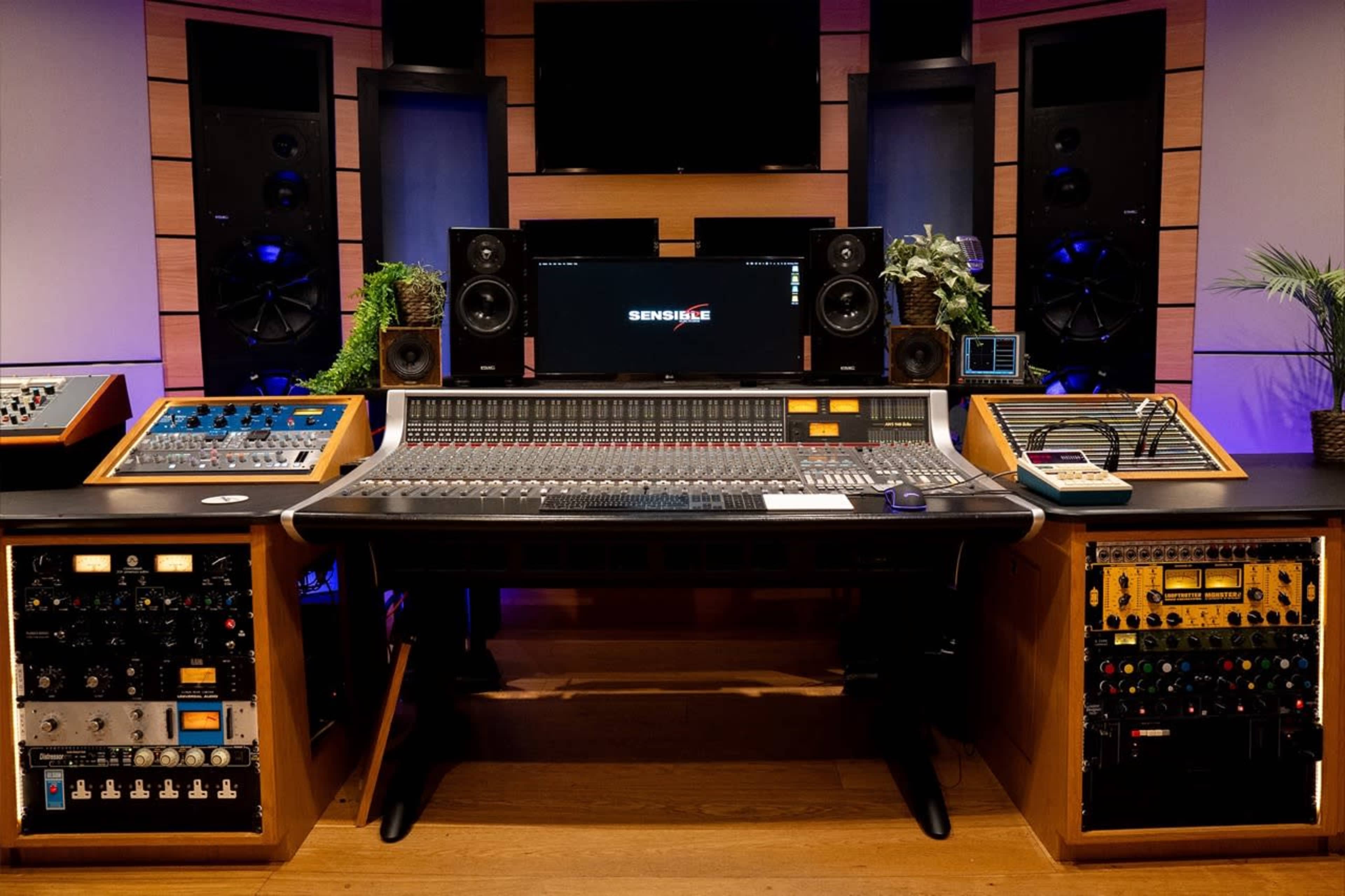 The image shows a professional music studio with a large mixing console at its center, flanked by audio equipment and speakers on either side.