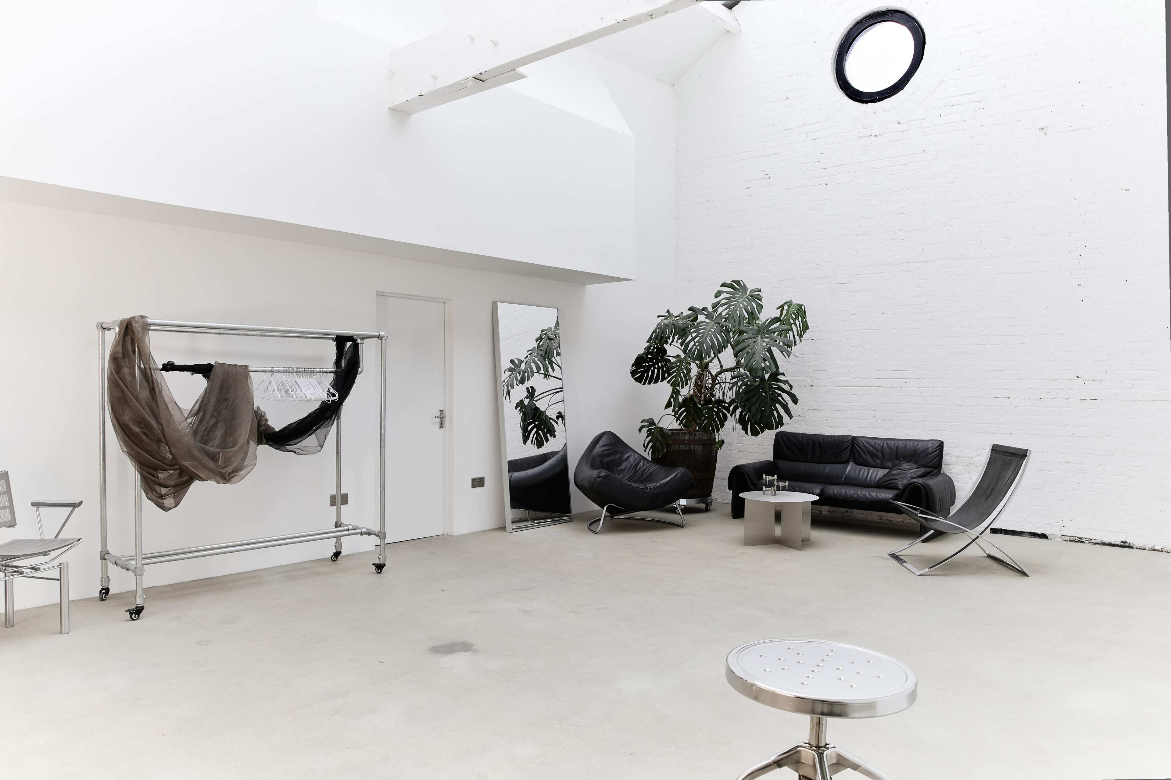 The image shows a spacious, minimalist interior with white walls, a seating area featuring a black couch and chairs, a clothing rack, and a large mirror.