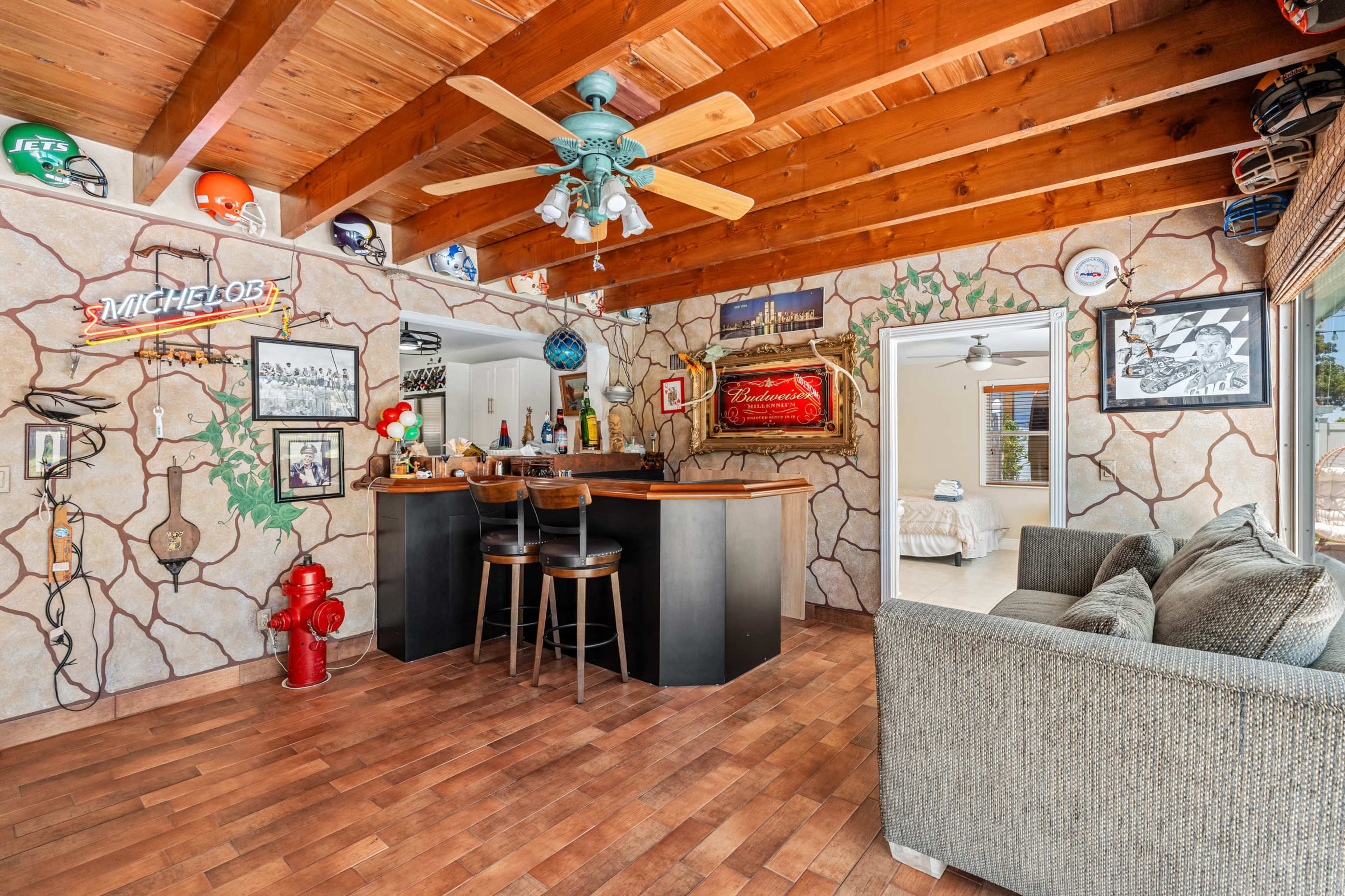 The image shows a cozy bar area with a wooden ceiling, featuring a few bar stools, a variety of decorations on the walls, and a small living space with a sofa in the foreground.