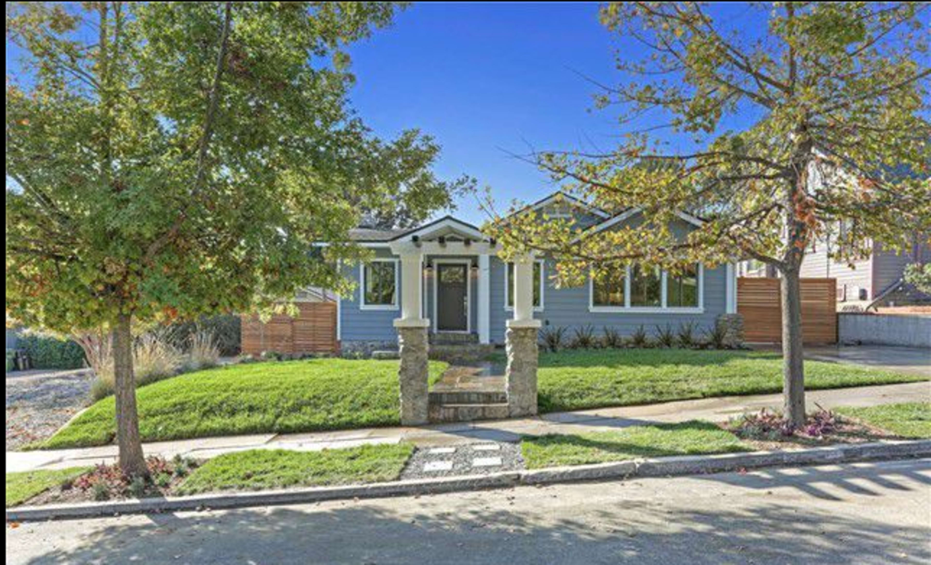 A single-story blue house with a stone porch is surrounded by green grass and trees, situated on a quiet street.