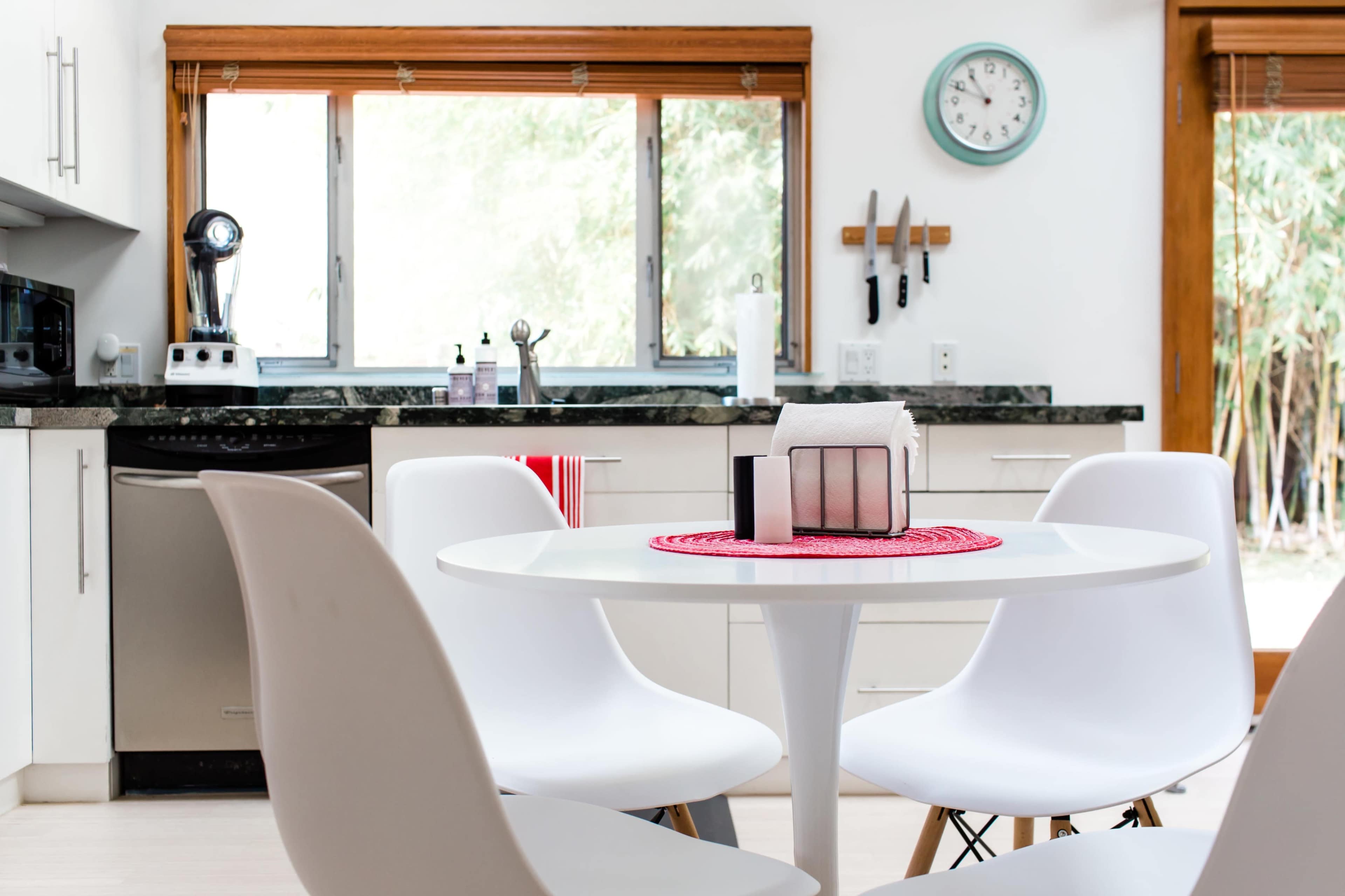 A modern kitchen features a round white table with four chairs, a marble countertop, and large windows allowing natural light.