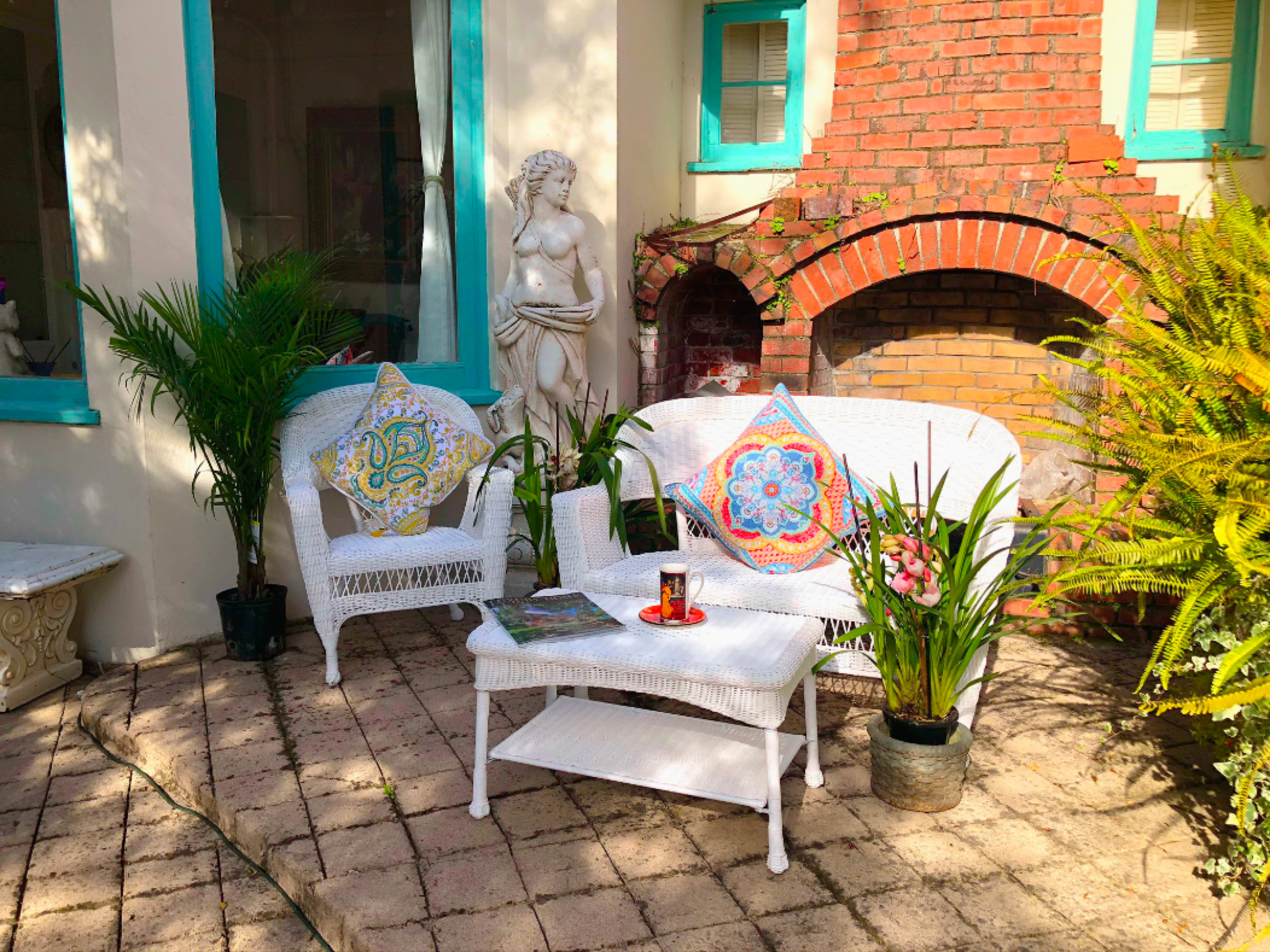 A cozy outdoor seating area features white wicker furniture, colorful cushions, a small table with a candle, and greenery around a brick wall with a sculpture.