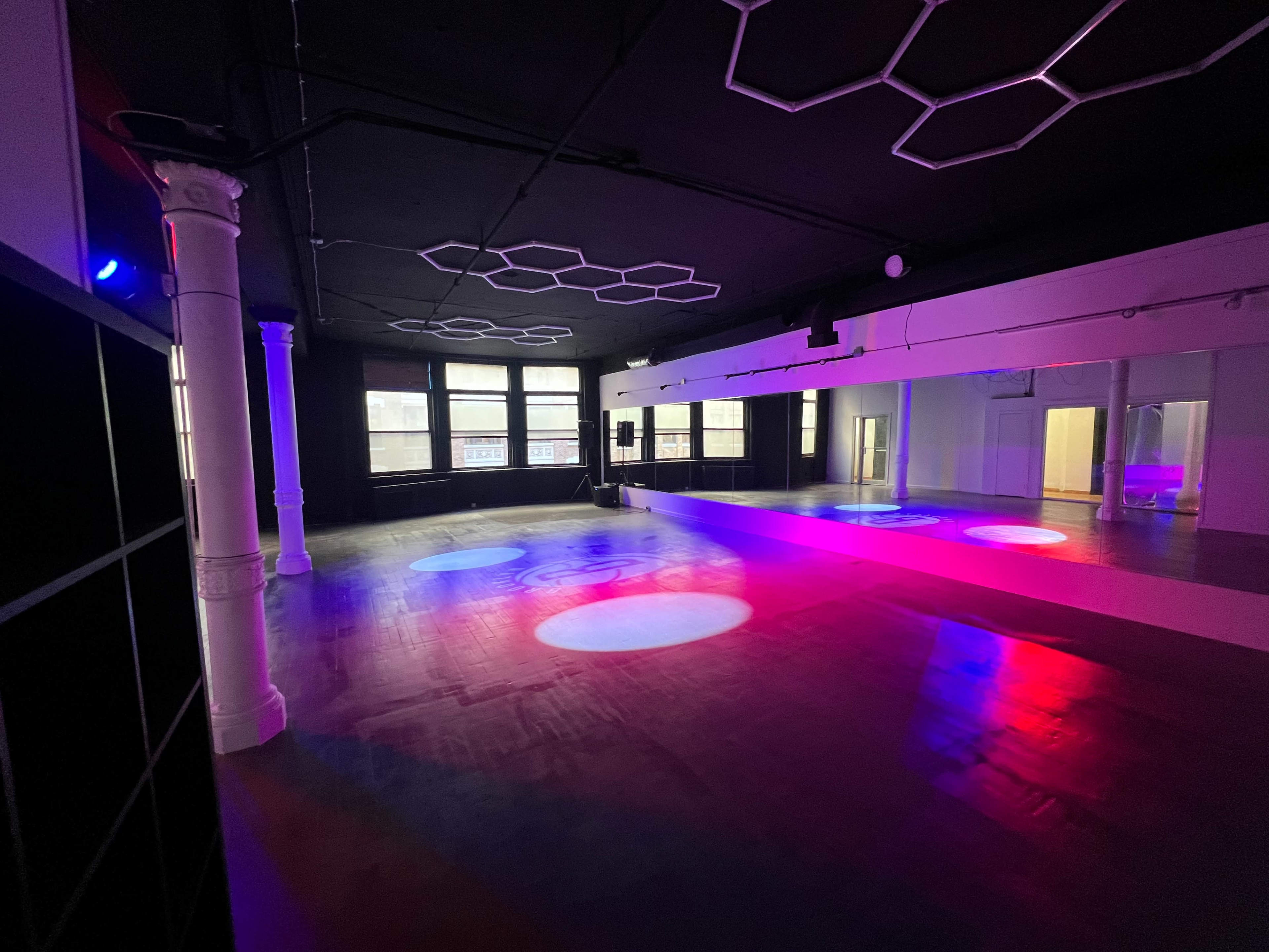 The image shows a spacious dance studio with a mirrored wall, wooden flooring, and color-changing lights illuminating the room.
