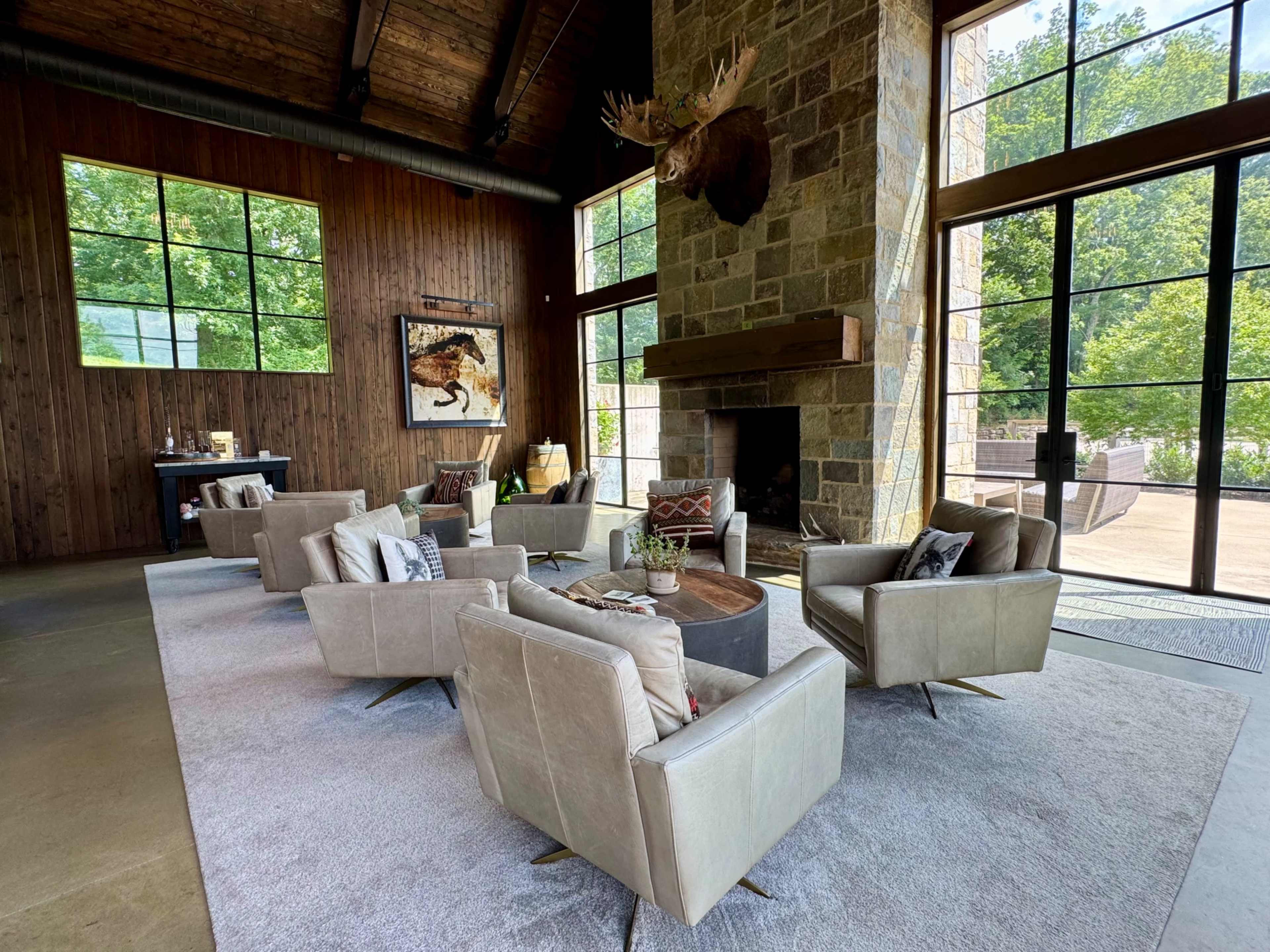The image shows a spacious indoor living area featuring a stone fireplace, large windows, and several beige armchairs arranged around a round coffee table on a light gray rug.