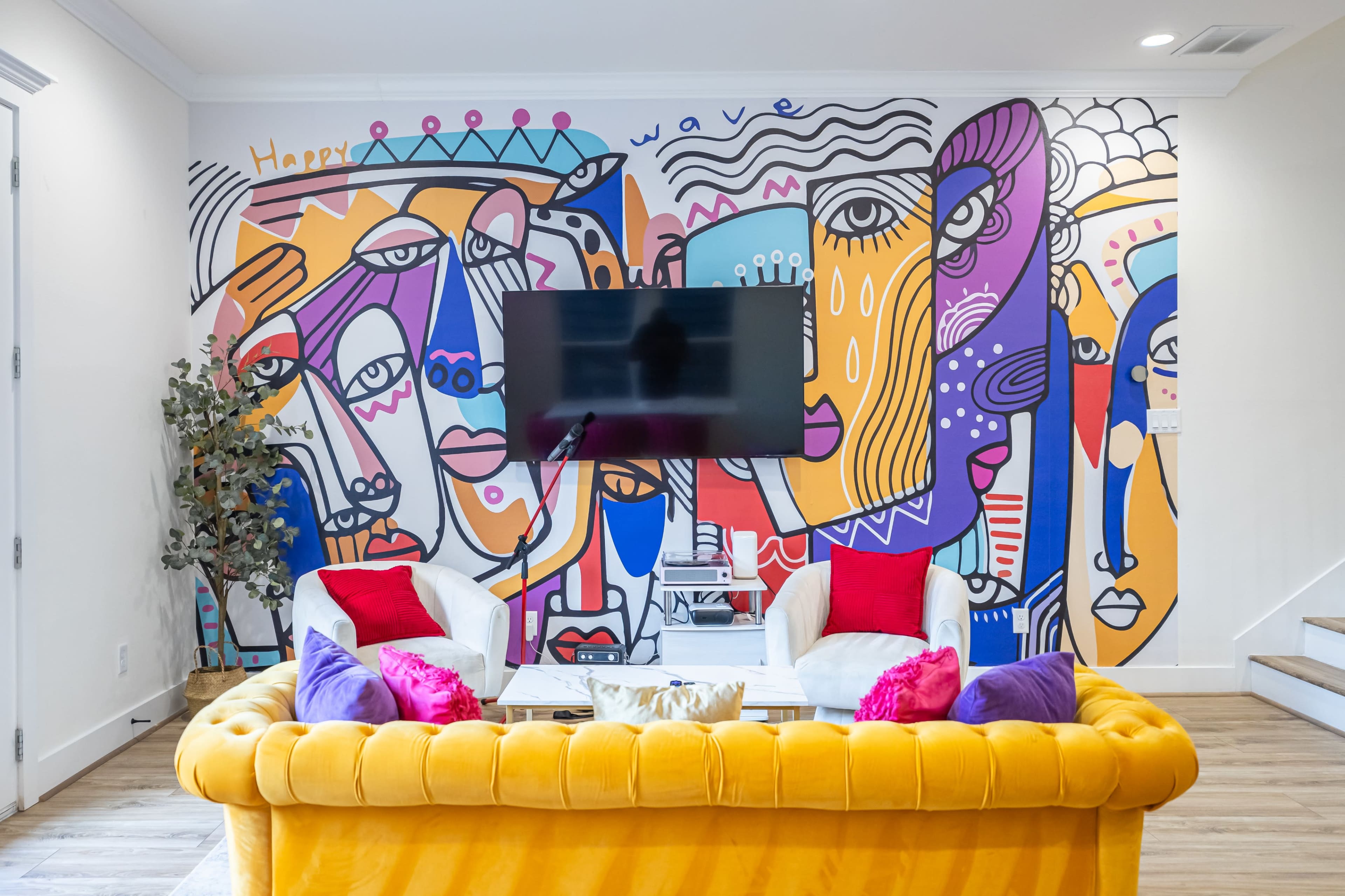 The image shows a brightly decorated living room with a large mural featuring abstract faces on the wall, a yellow sofa, and a television mounted above a white coffee table.