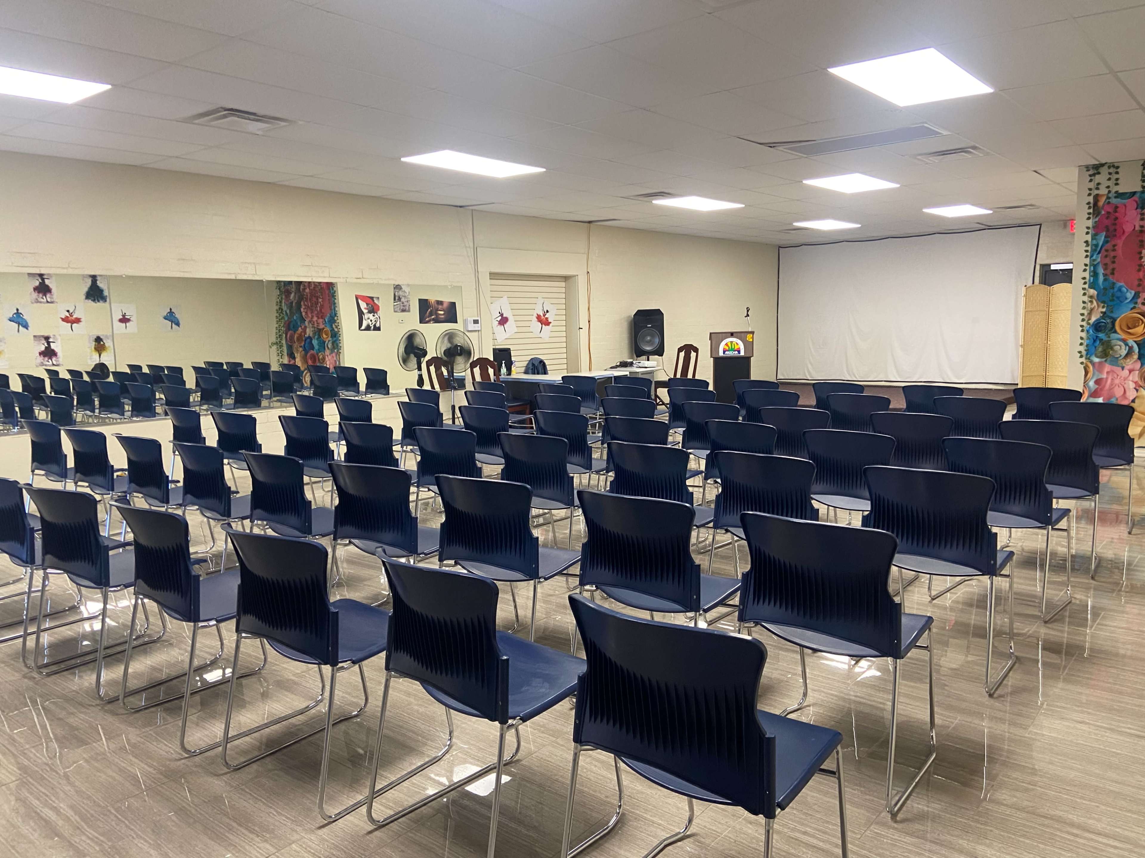 A classroom or meeting space filled with rows of blue chairs facing a blank projector screen.