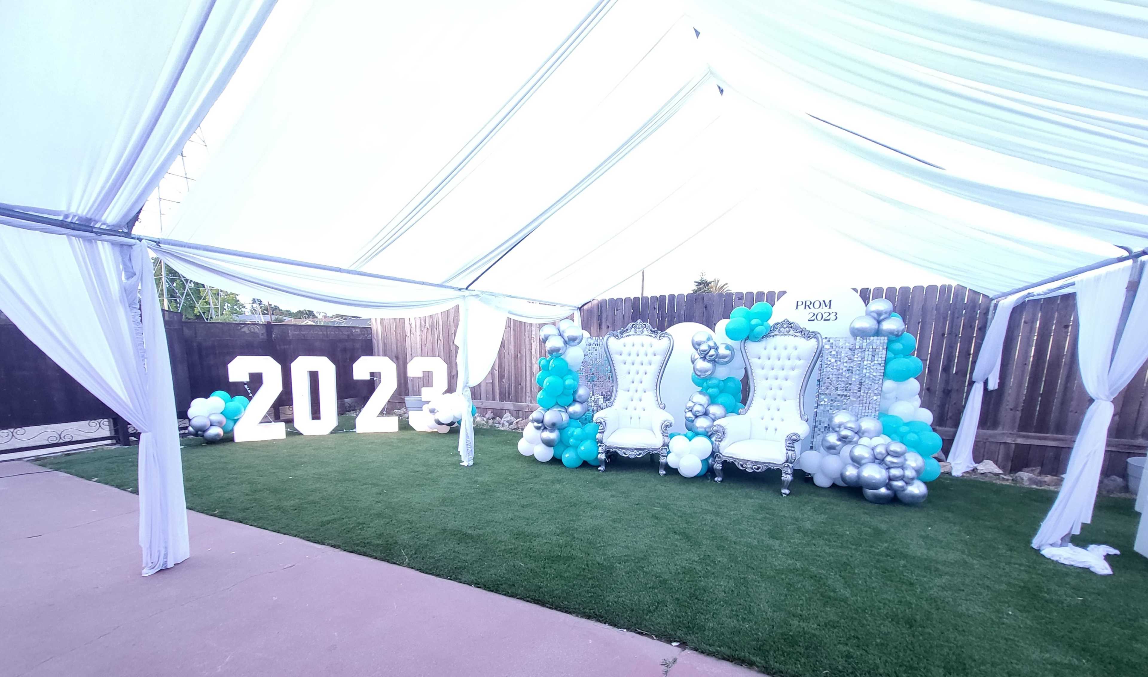 A decorated area for a prom event featuring large "2023" numbers, two white throne chairs, and an arrangement of balloons in teal, silver, and white.