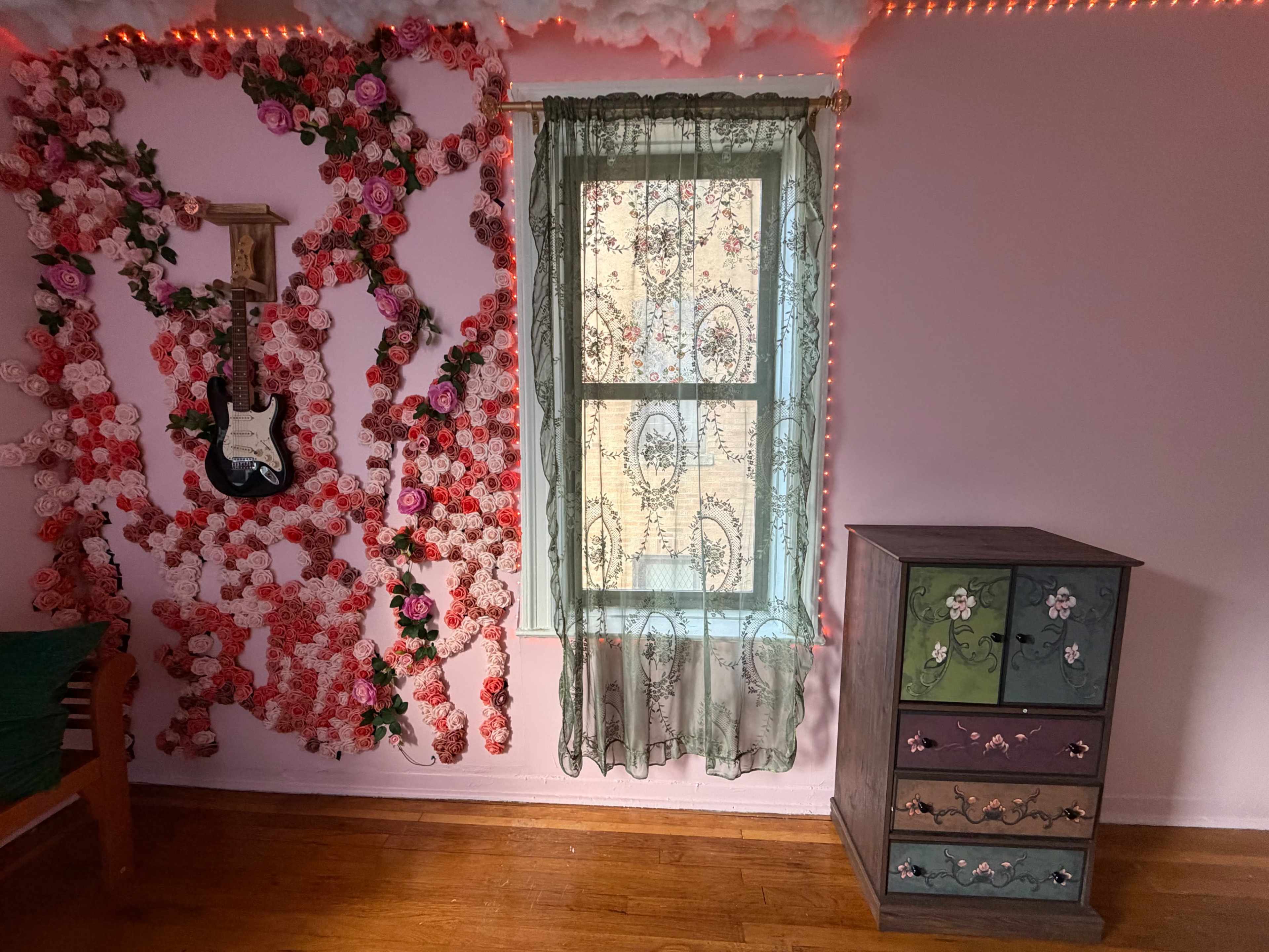 The image shows a room with a floral wall decoration, a window covered by a sheer curtain, and a small dresser with colored drawers.