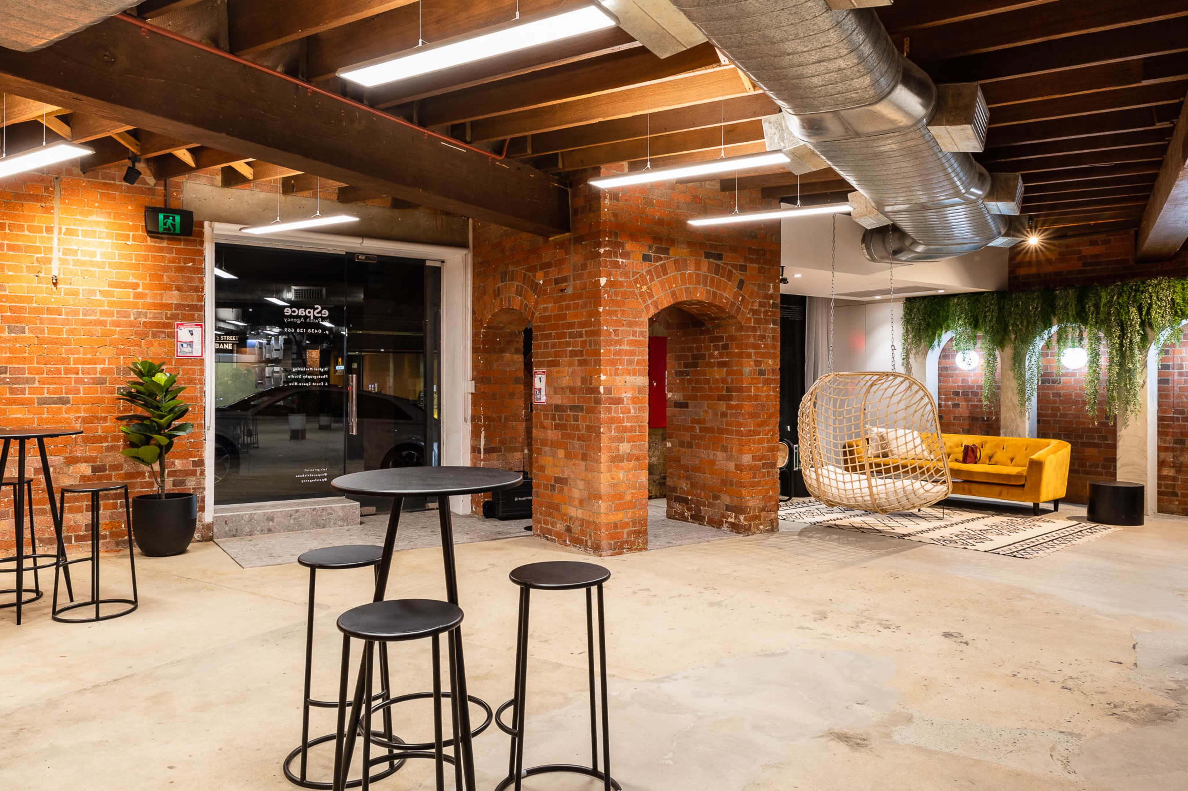 The interior of a modern space features exposed brick walls, industrial lighting, a seating area with a yellow couch, and a decorative hanging chair.