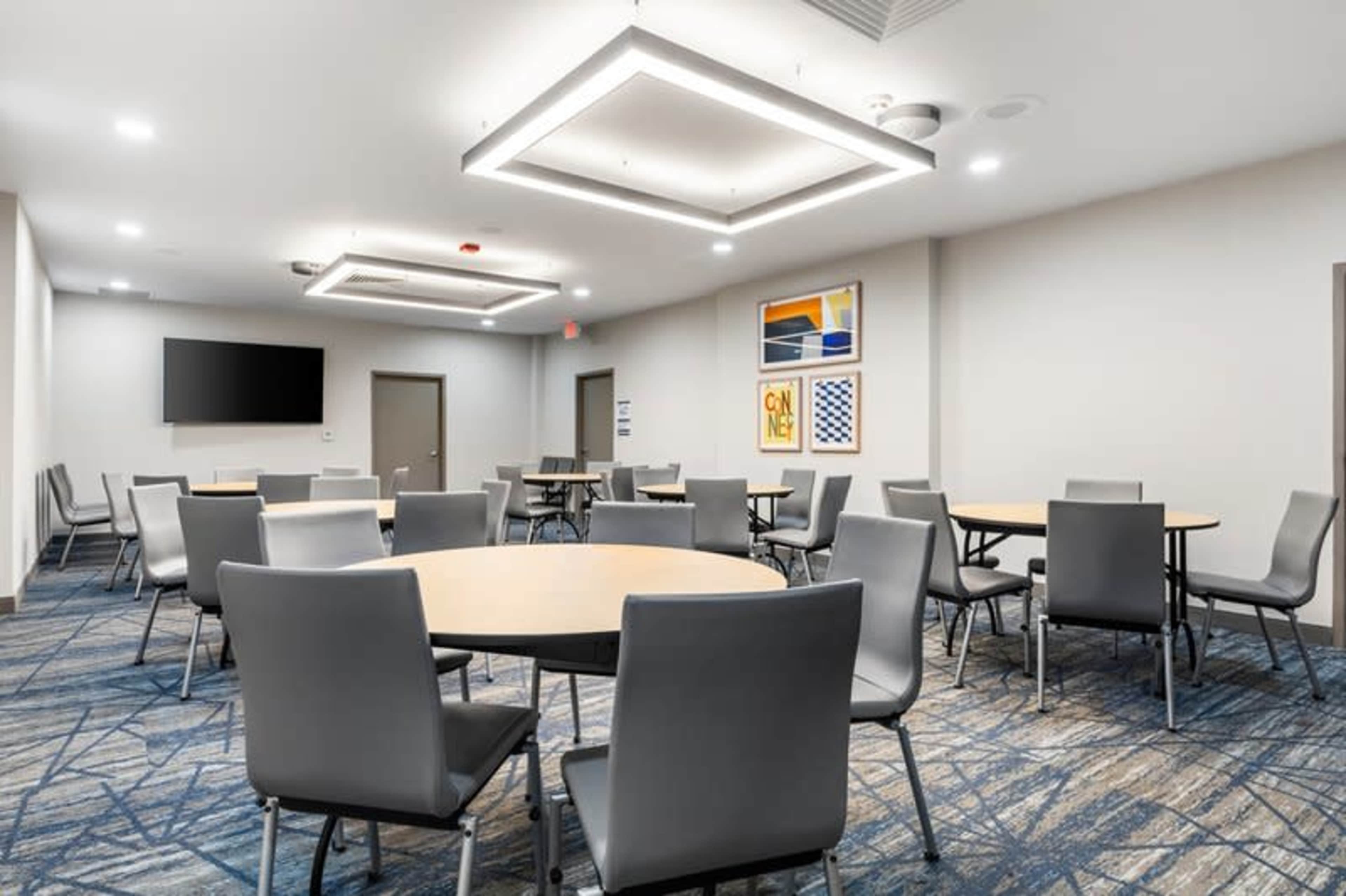 The image shows a modern conference room with several round tables and chairs, illuminated by rectangular ceiling lights.