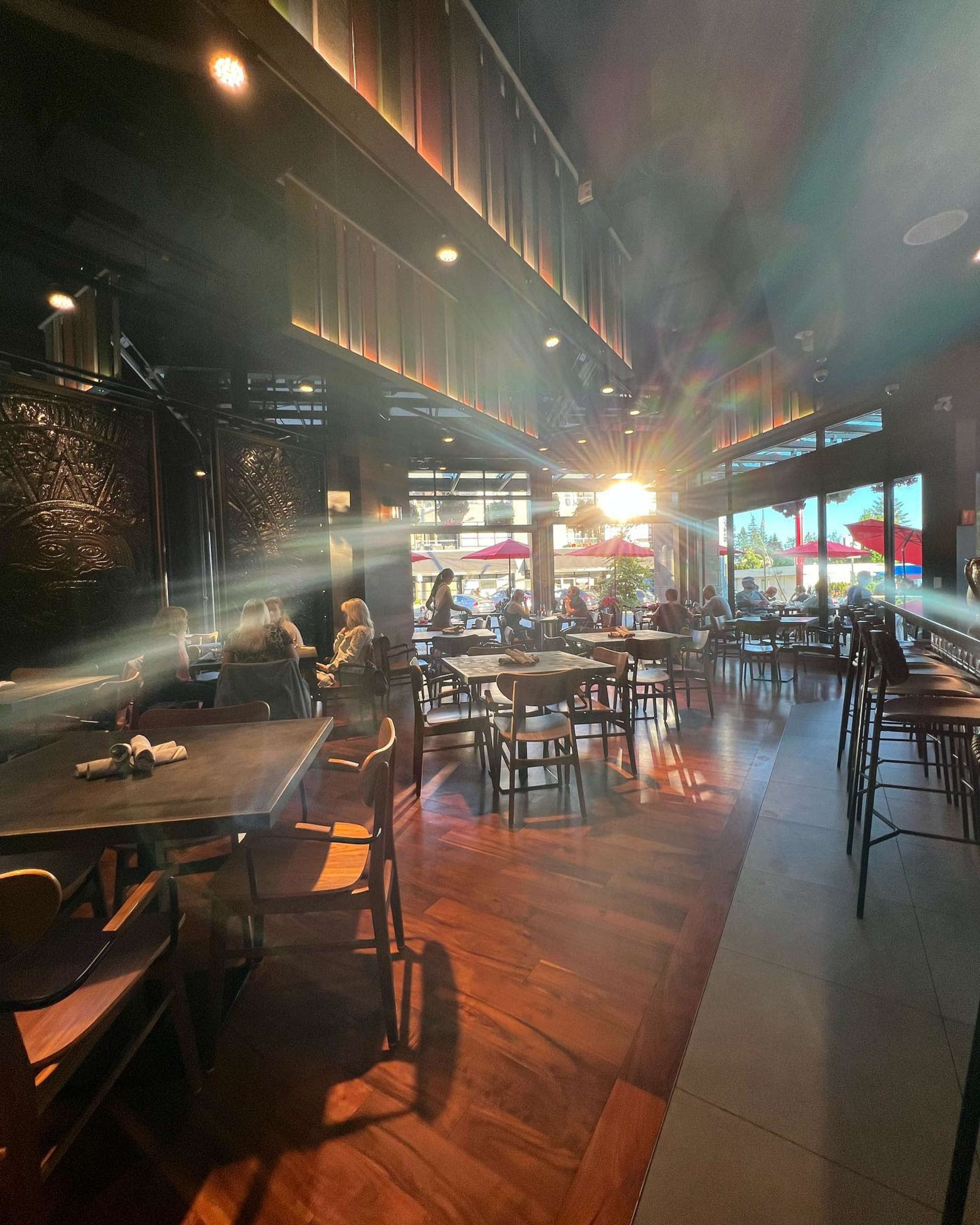 The interior of a modern restaurant features wooden tables and chairs with sunlight streaming through large windows.