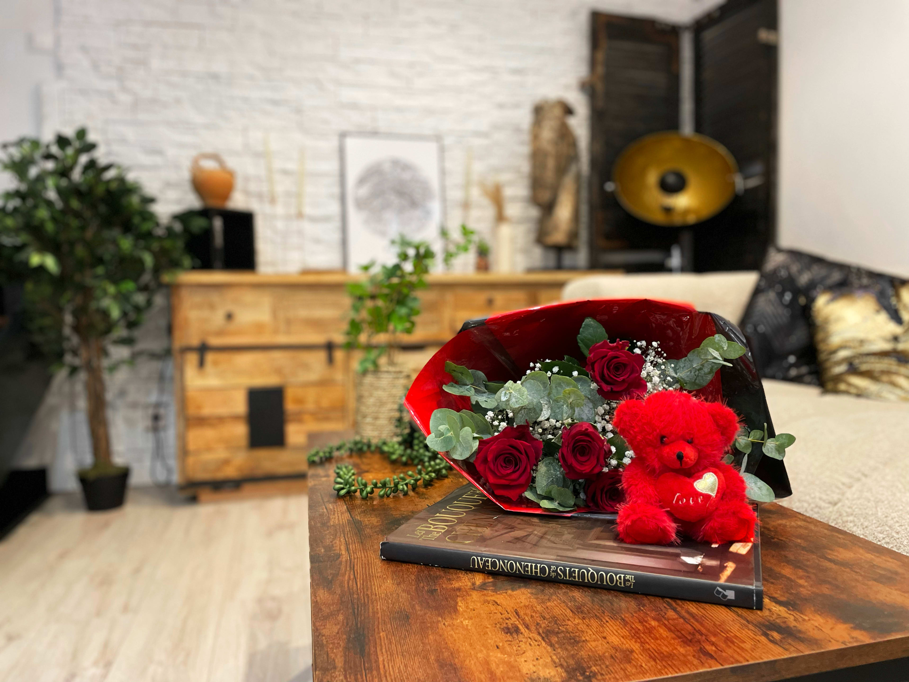 A bouquet of red roses and a stuffed bear sit on a wooden coffee table in a cozy living room with decorative plants and furniture.