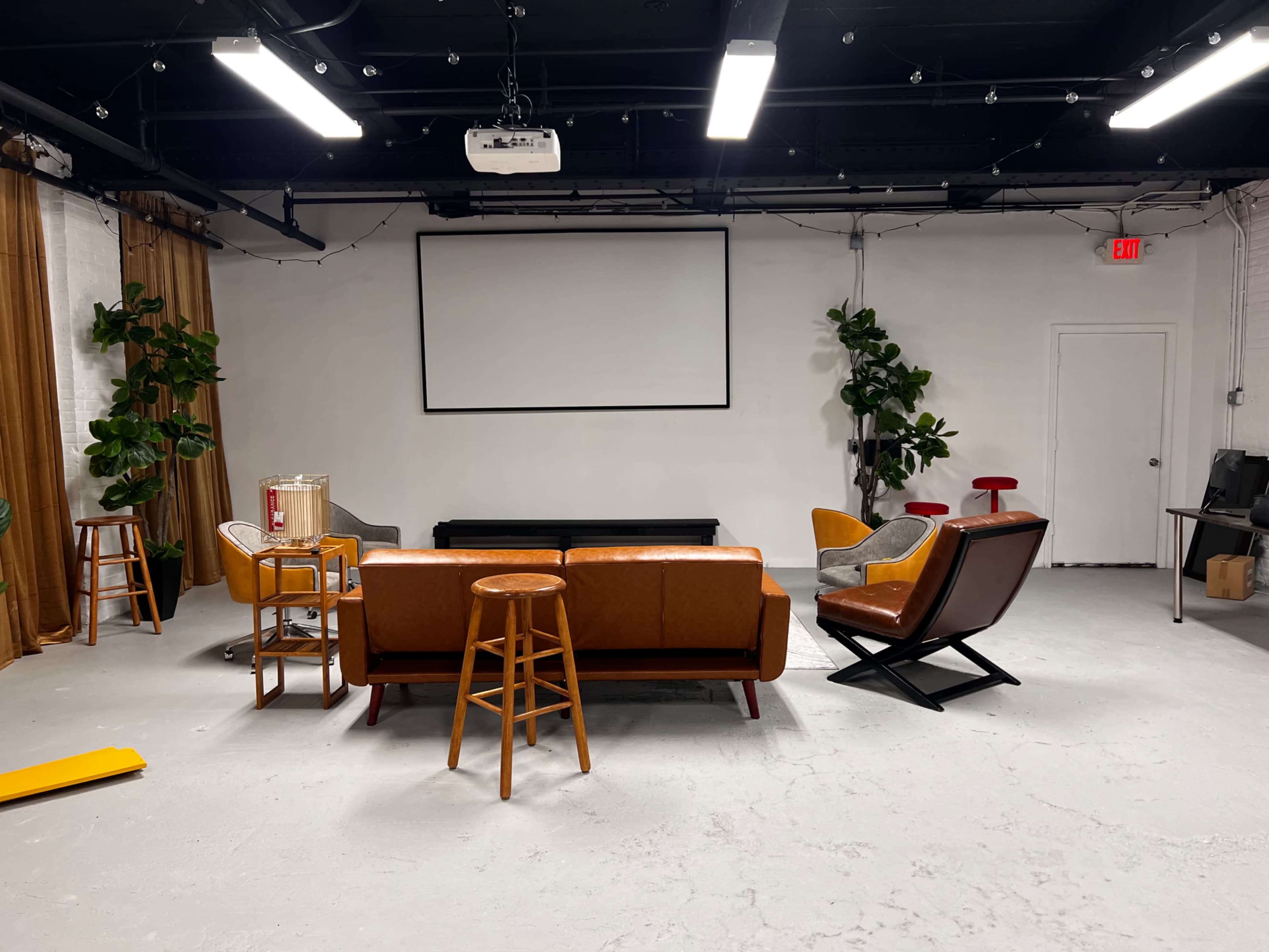 A spacious room with a projector screen, featuring a brown leather couch, several chairs, two wooden stools, and decorative plants.