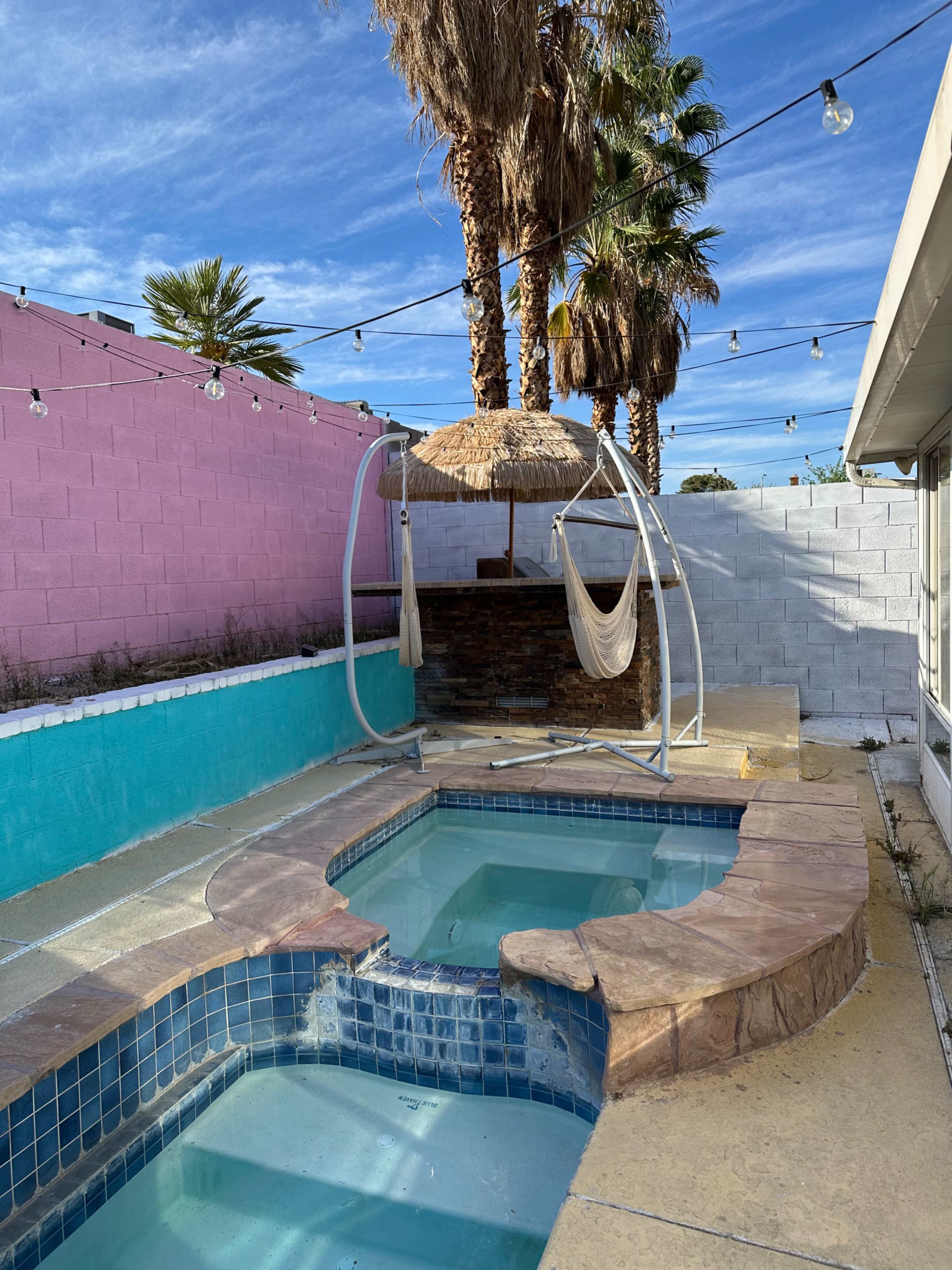 The image shows a small pool area featuring a blue-tiled spa and a larger pool, surrounded by palm trees and a pink and white wall, with a thatched umbrella and hammock setup nearby.