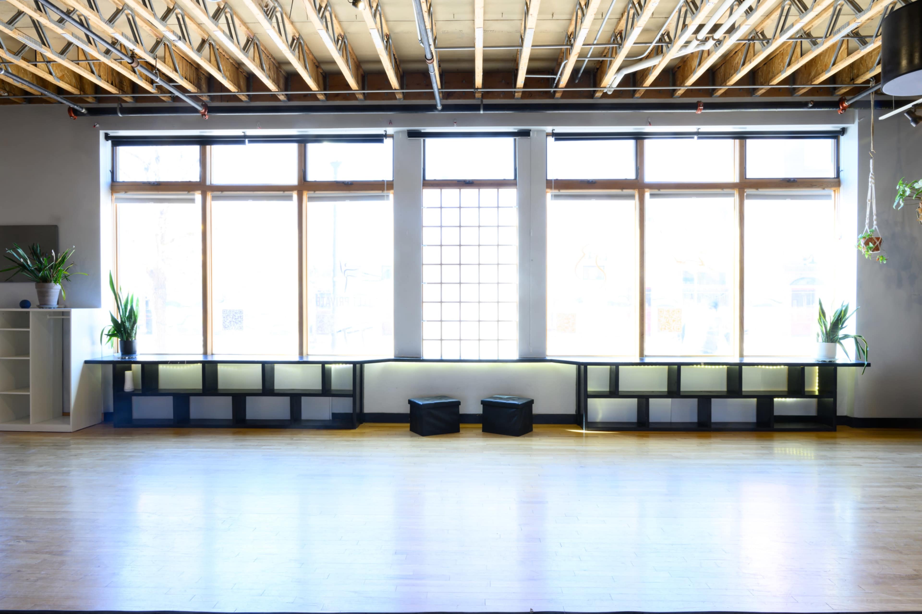 The image shows a spacious, well-lit room featuring large windows, a wooden ceiling, and a minimalist shelf along the walls.