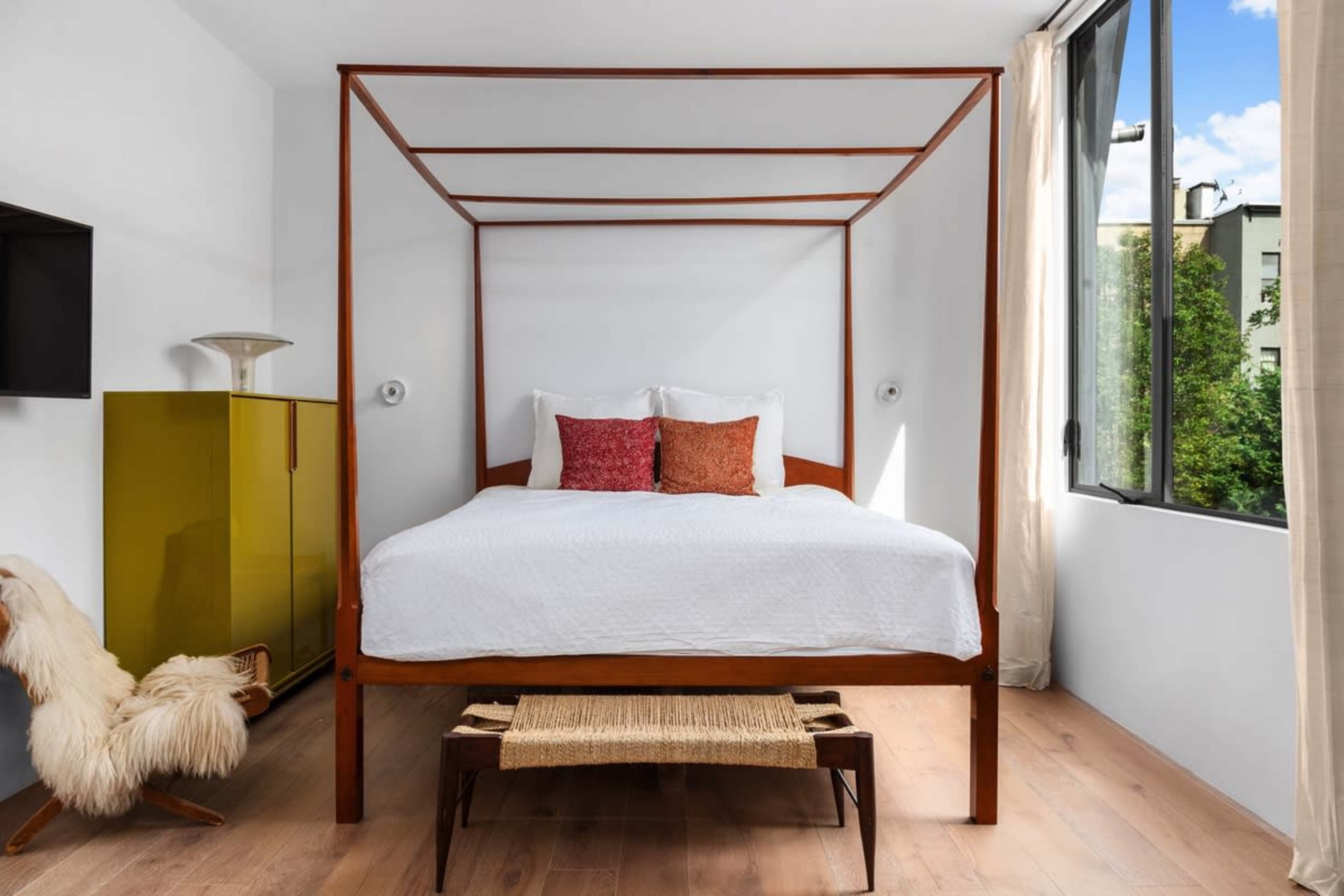 A four-poster bed with white bedding is positioned in a bright room featuring large windows and a yellow cabinet.