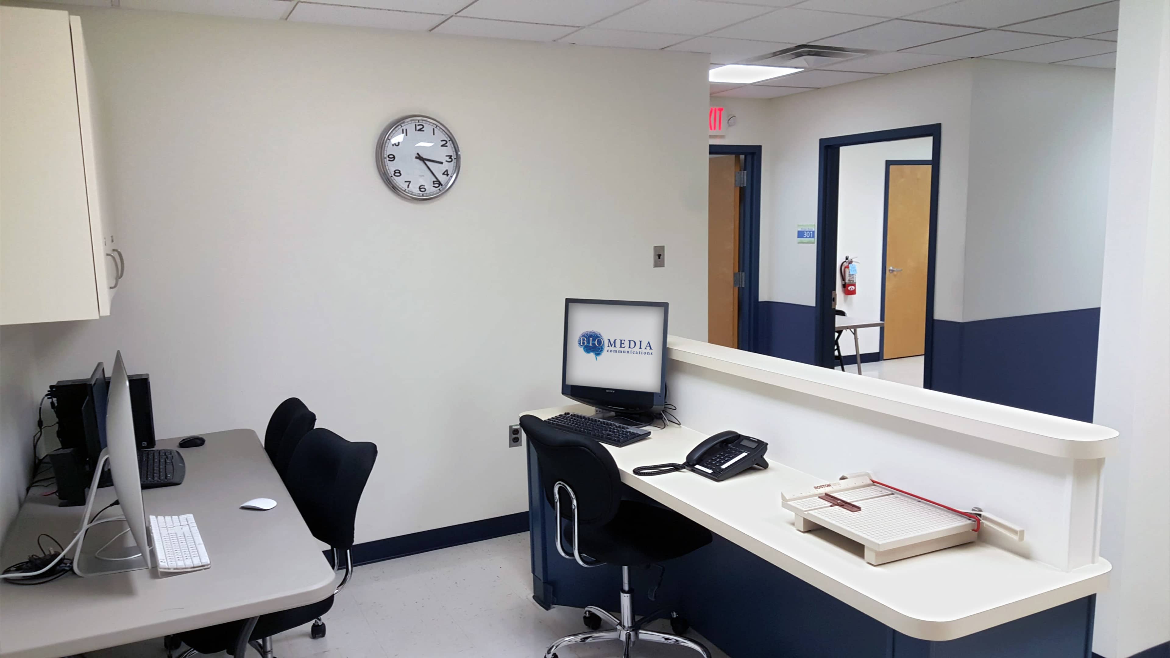A neatly arranged office space featuring a wall clock, computer stations, a telephone, and a receptionist desk with a wooden registration board.