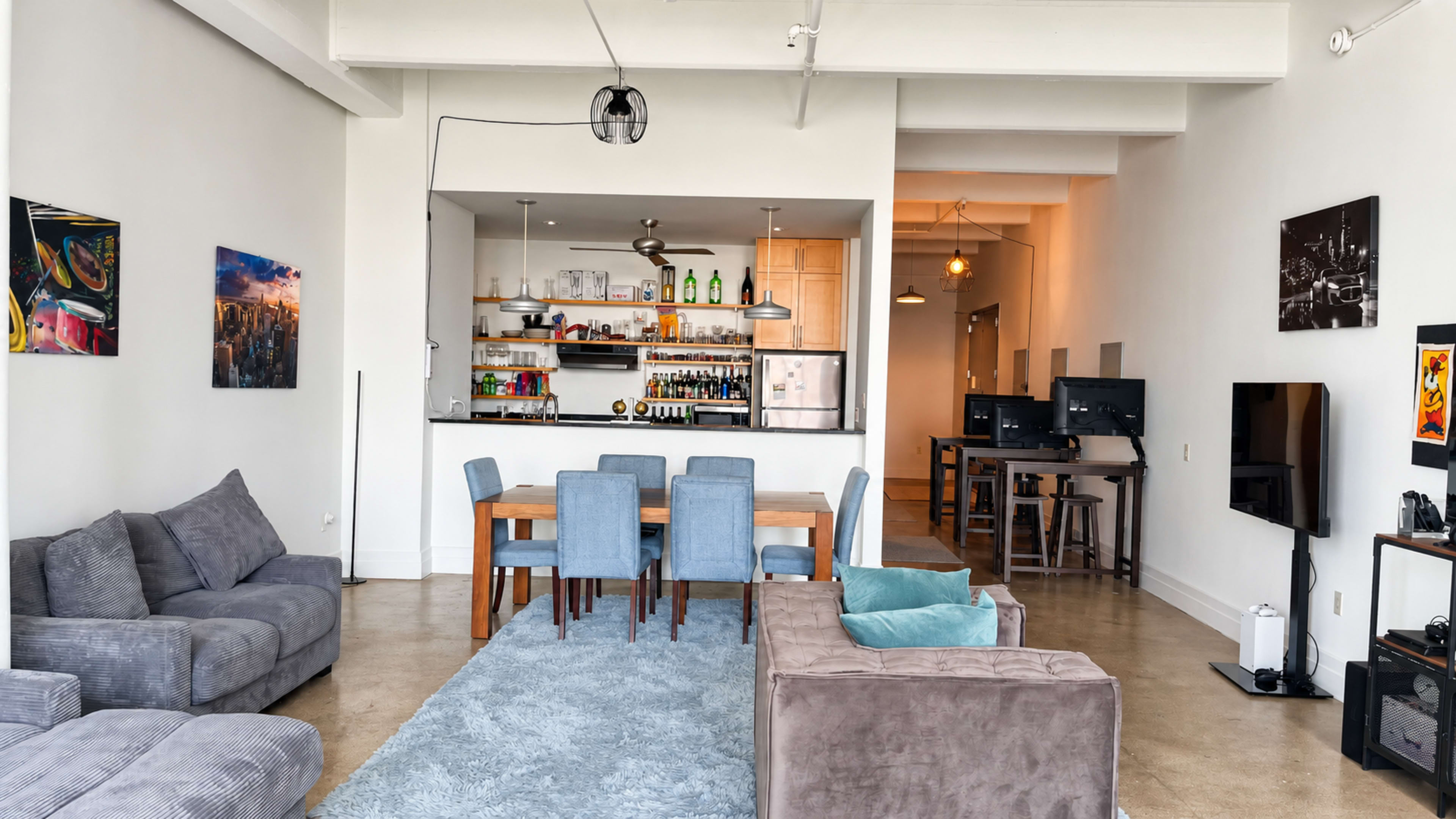 The image shows a modern, open-concept living space featuring a seating area with gray sofas, a dining table with blue chairs, and a kitchen in the background with shelves displaying various items.