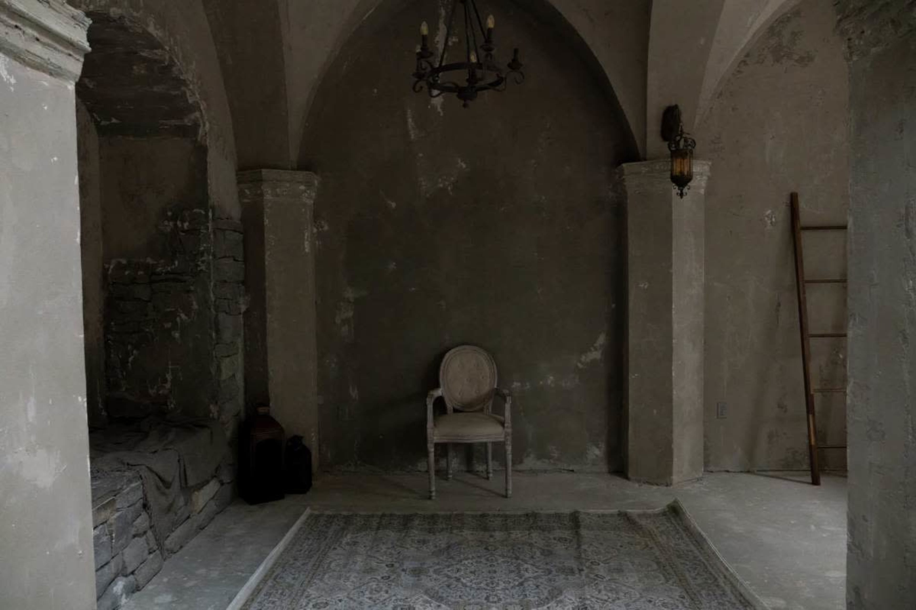 An empty room with stone walls, a lone chair in the center, and a rug on the floor, illuminated by a chandelier and wall sconces.