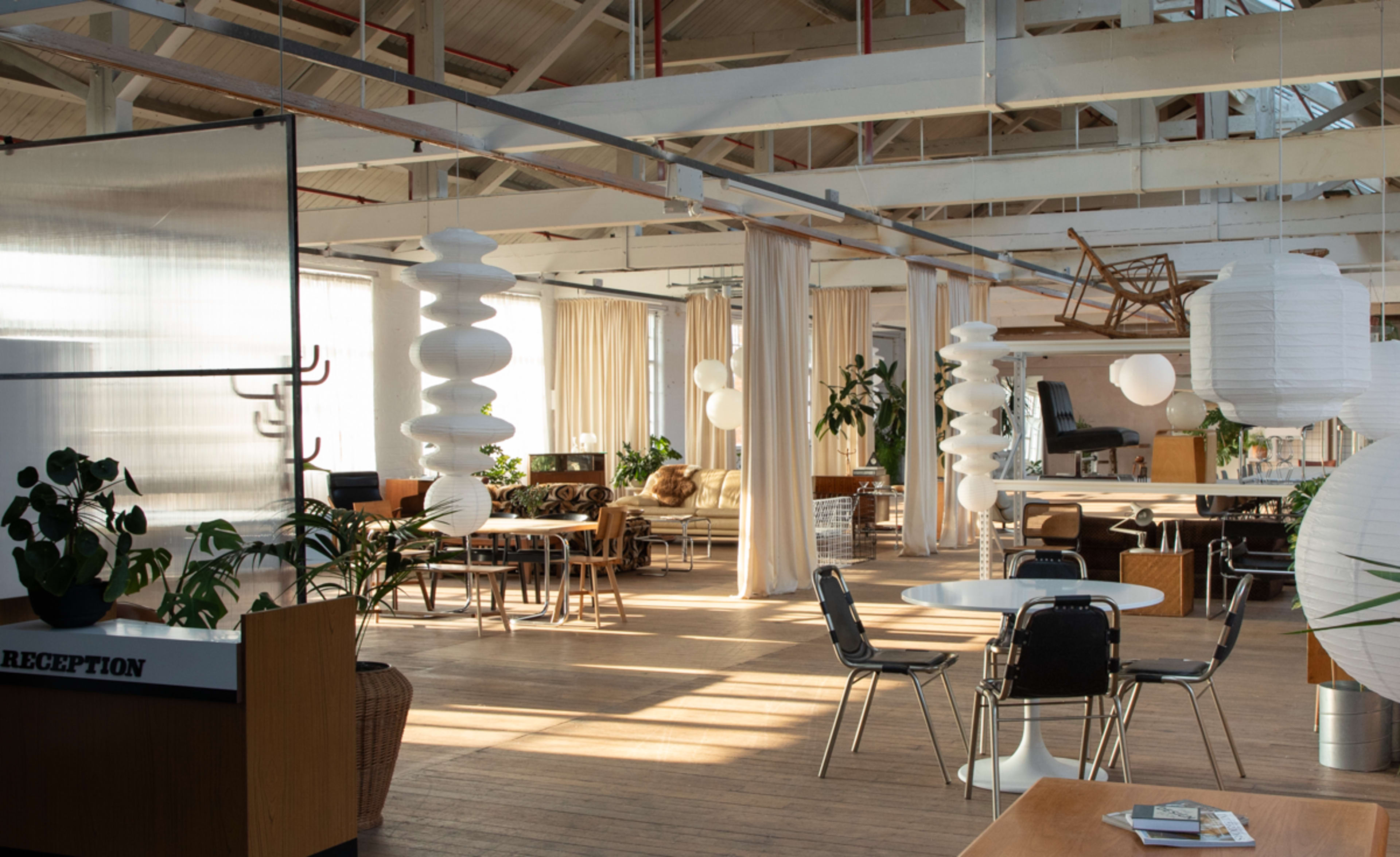 The image shows a spacious, modern interior with wooden flooring, plants, and a mix of tables and seating arranged around paper lanterns and curtains.