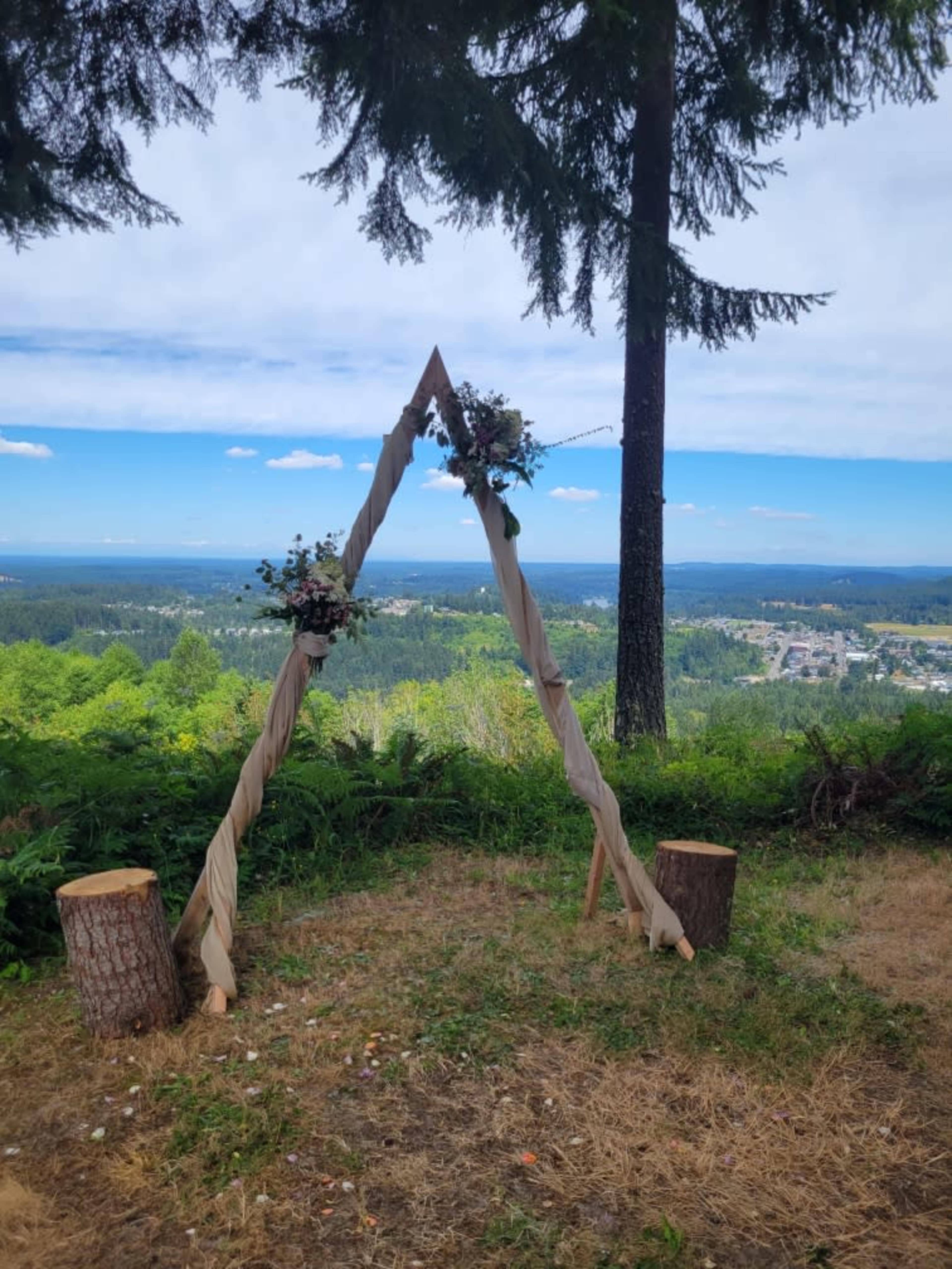 A triangular wooden arch adorned with floral arrangements stands on a grassy area overlooking a valley and distant hills under a blue sky.