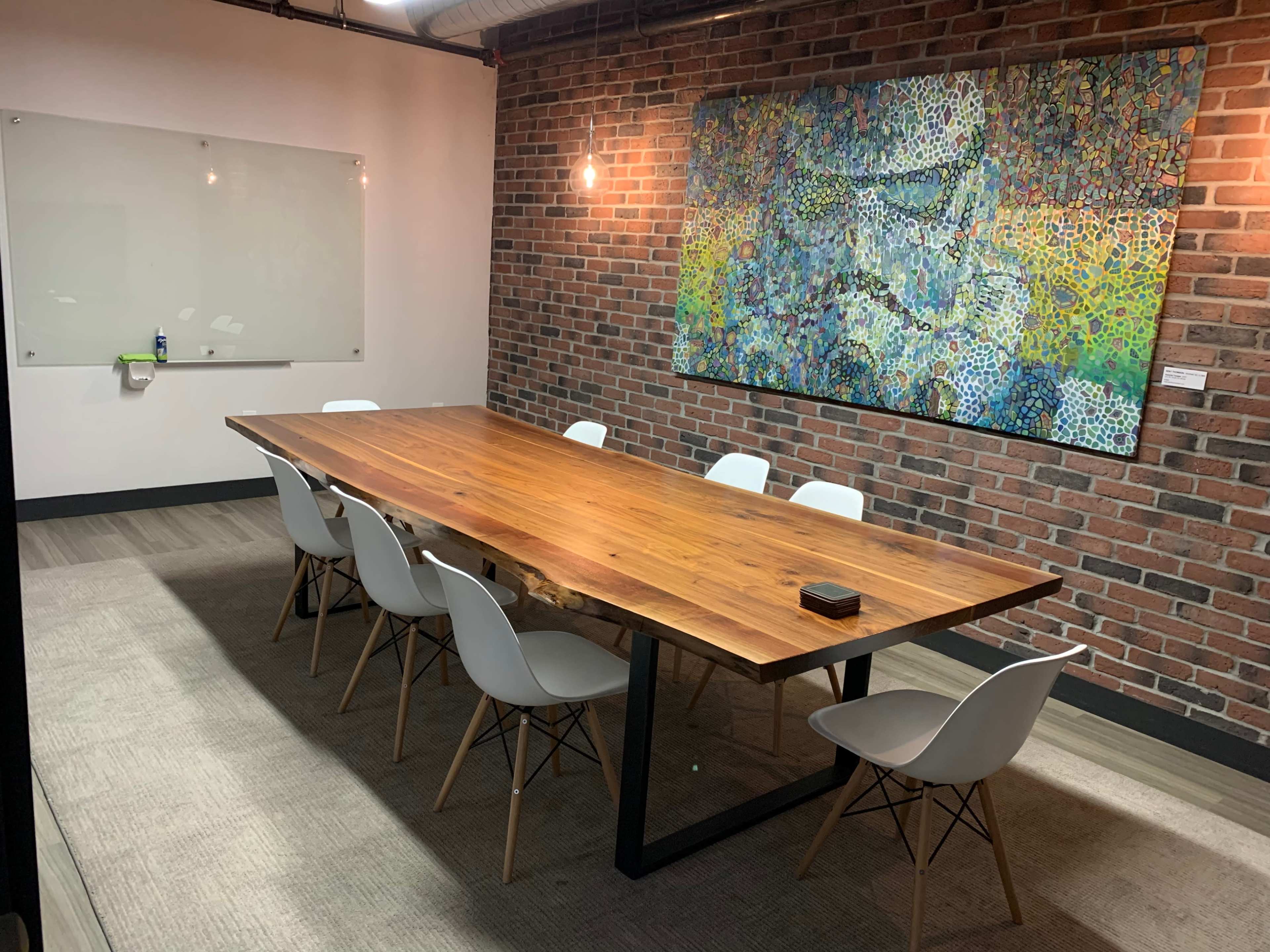 A conference room features a long wooden table surrounded by white chairs, with a colorful artwork displayed on a brick wall.