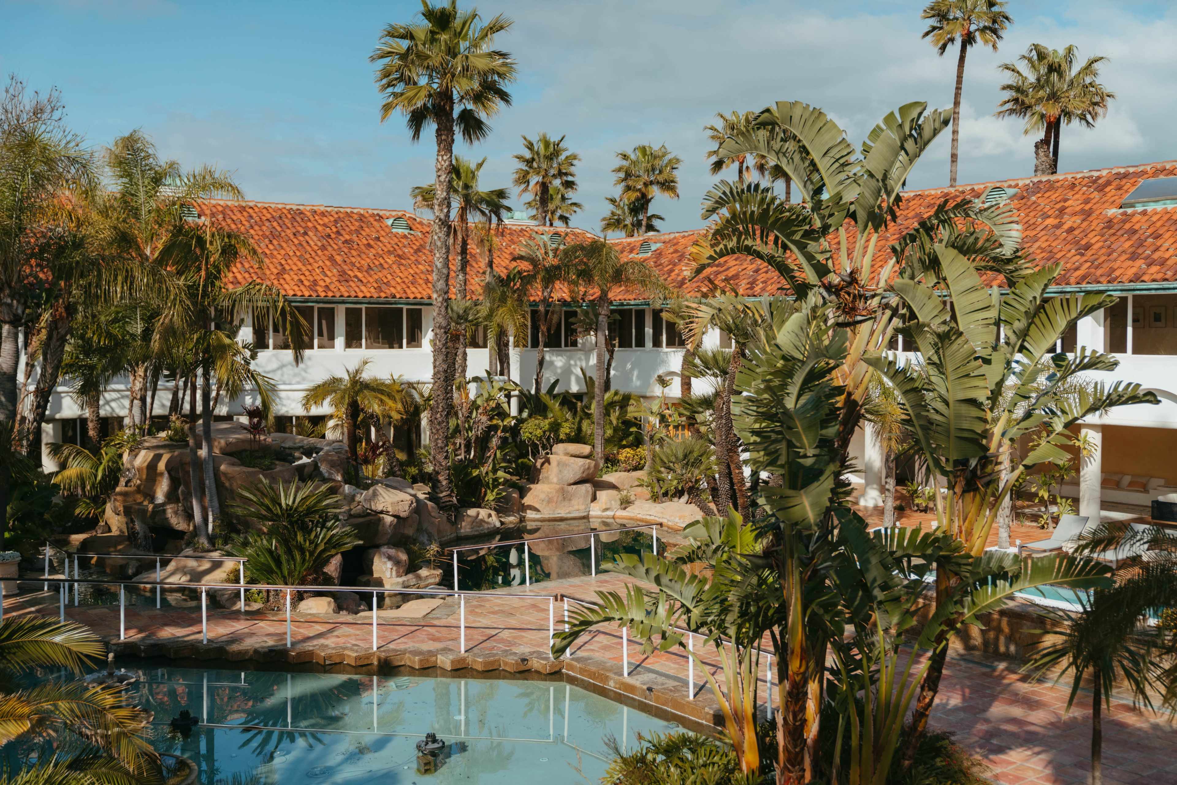 A tropical resort features a pool surrounded by palm trees and rock formations, with a building in the background.