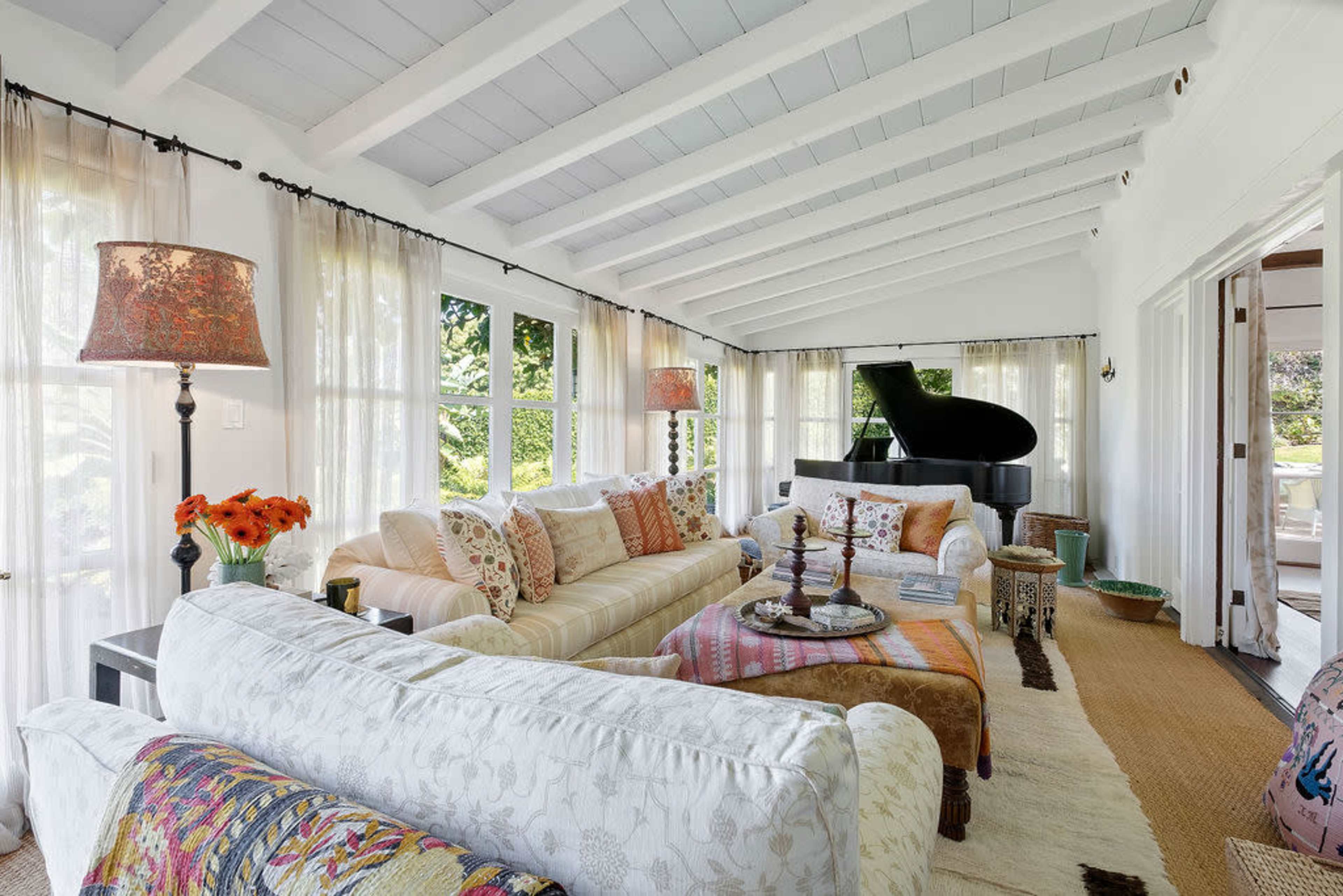 A sunlit living room features white-beamed ceilings, large windows with sheer curtains, and a grand piano in the background, surrounded by comfortable seating and a decorative rug.