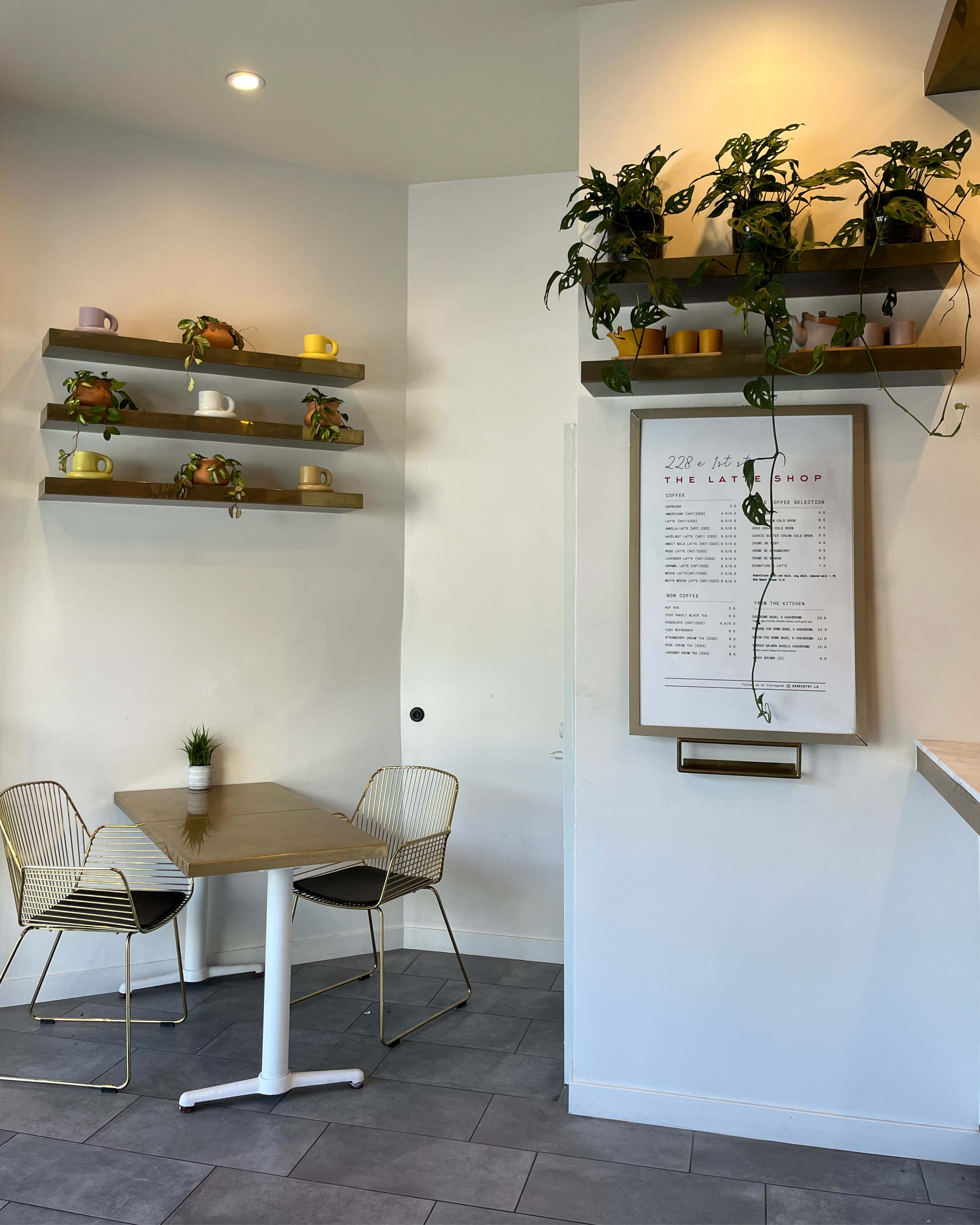A small café interior features a table with two wire chairs, shelves with colorful mugs and plants, and a menu display on the wall.