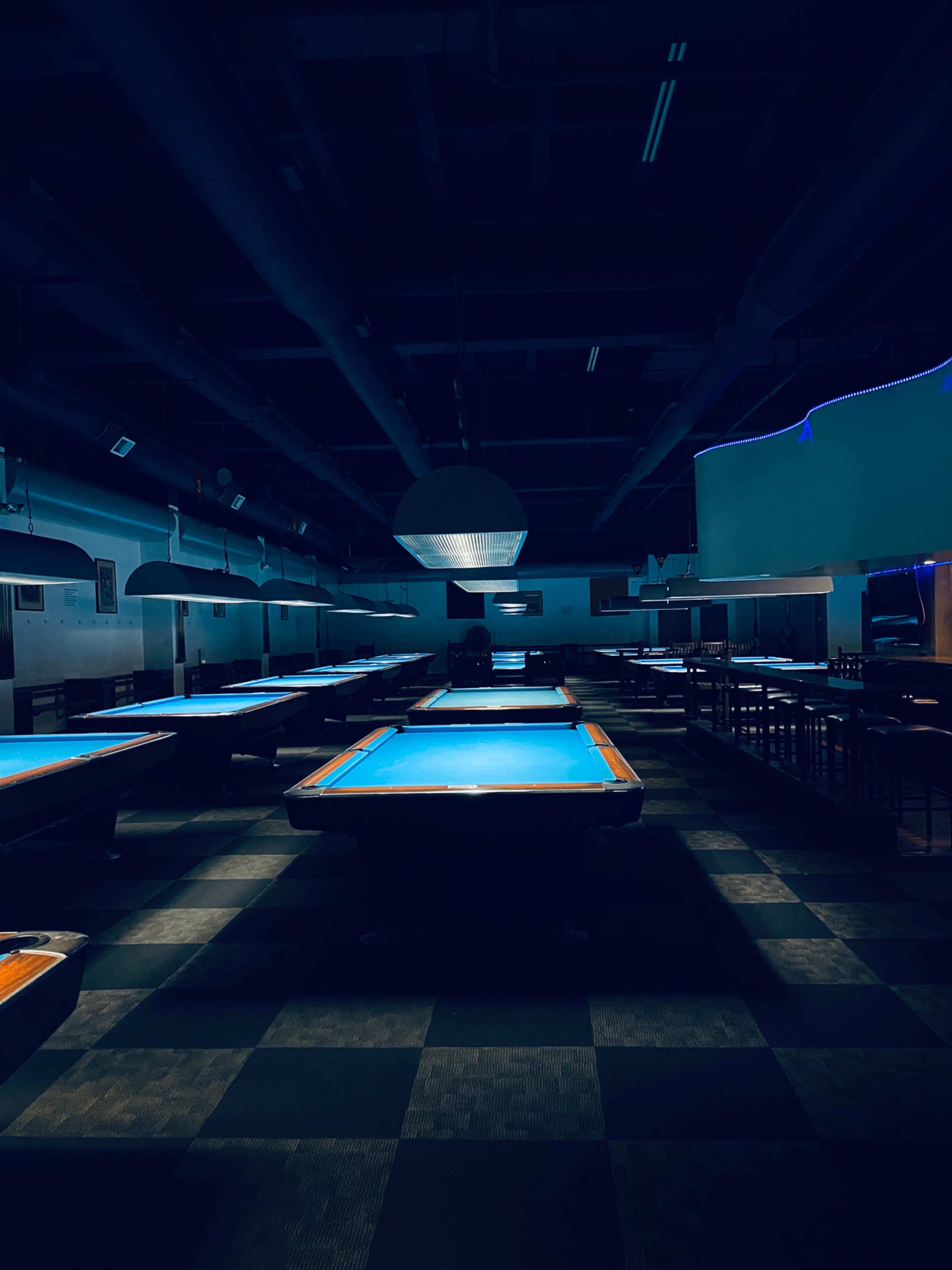 An empty pool hall features several tables, each illuminated by overhead lights, under dim conditions.