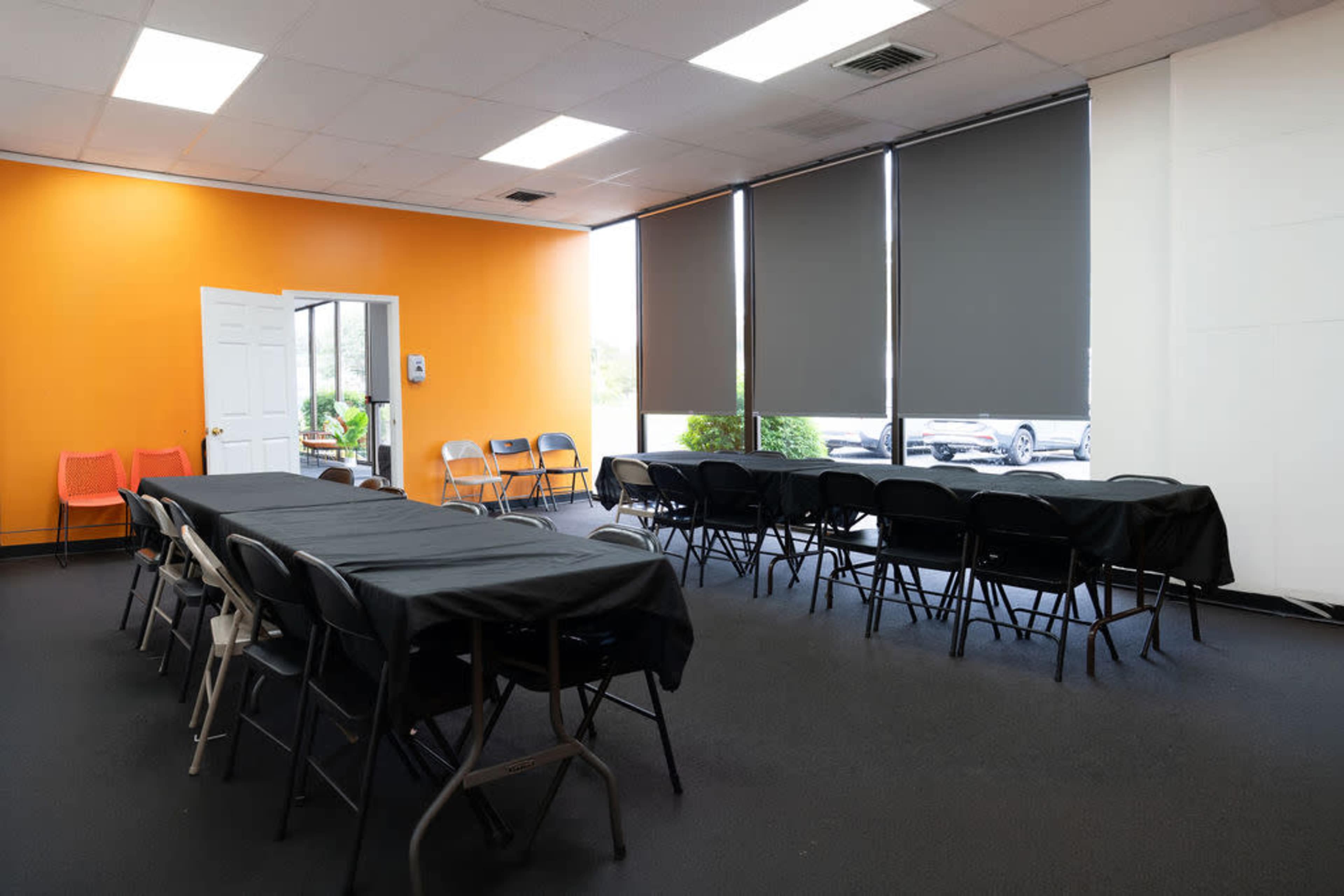 The image shows a meeting room with tables covered in black cloth, arranged with black chairs, against an orange wall and large windows with dark blinds.