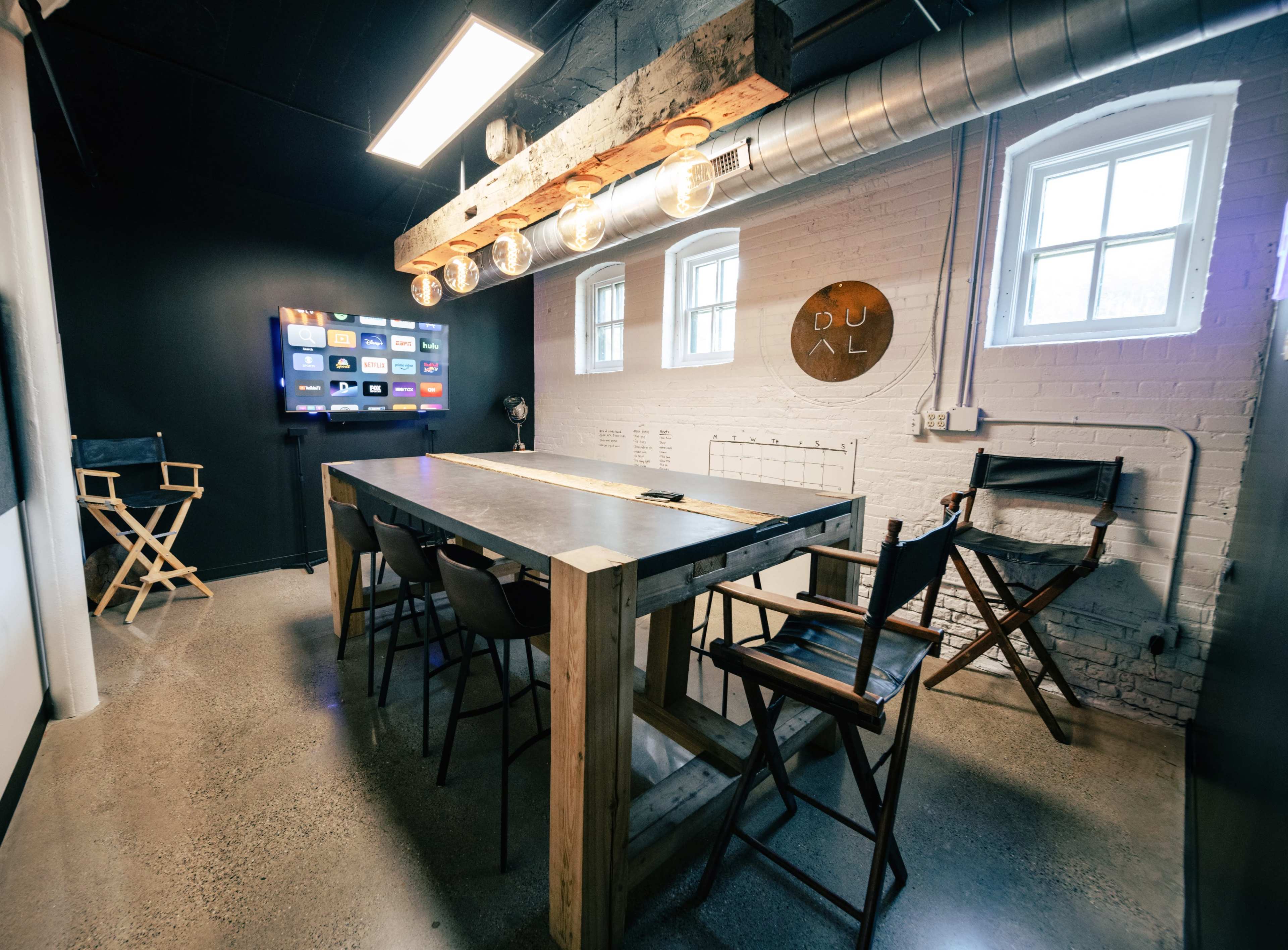 The image shows a modern meeting room with a long wooden table, high chairs, and a television screen mounted on the wall.