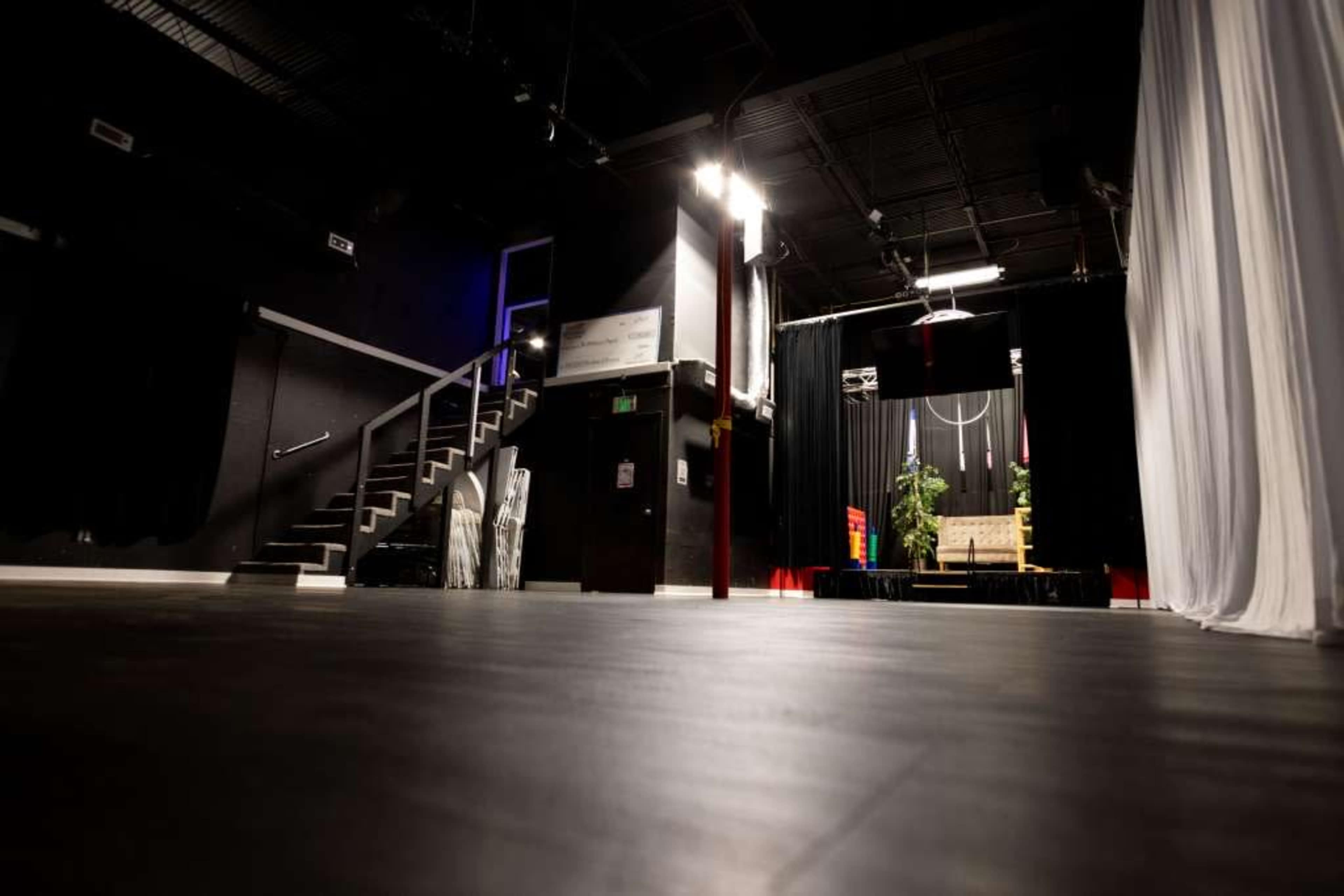 The image shows a dimly lit performance space with a stage at the end, featuring a chair and plants, and a staircase leading to the upper level on the left.