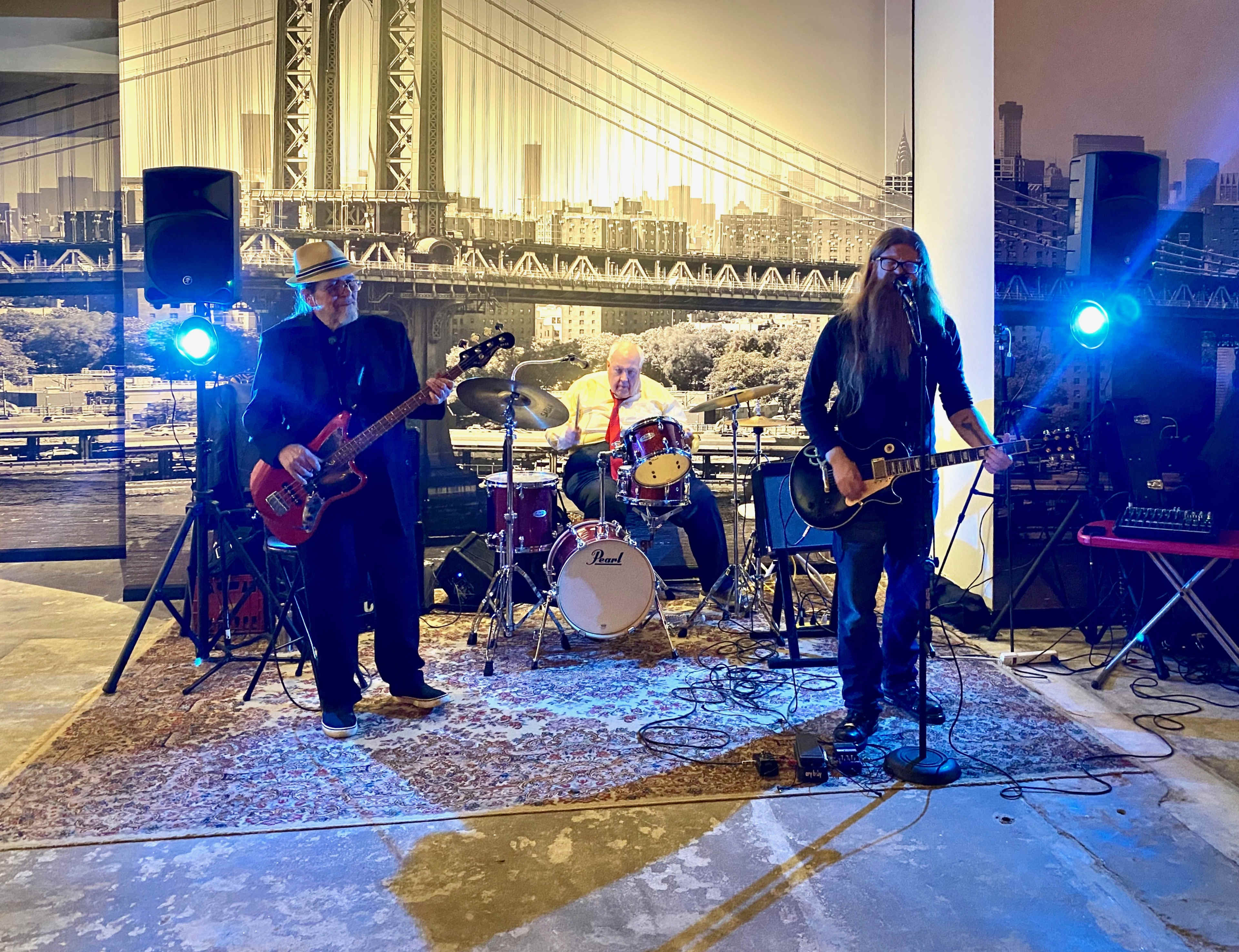 A band performs on a stage with instruments, set against a backdrop of the Brooklyn Bridge.