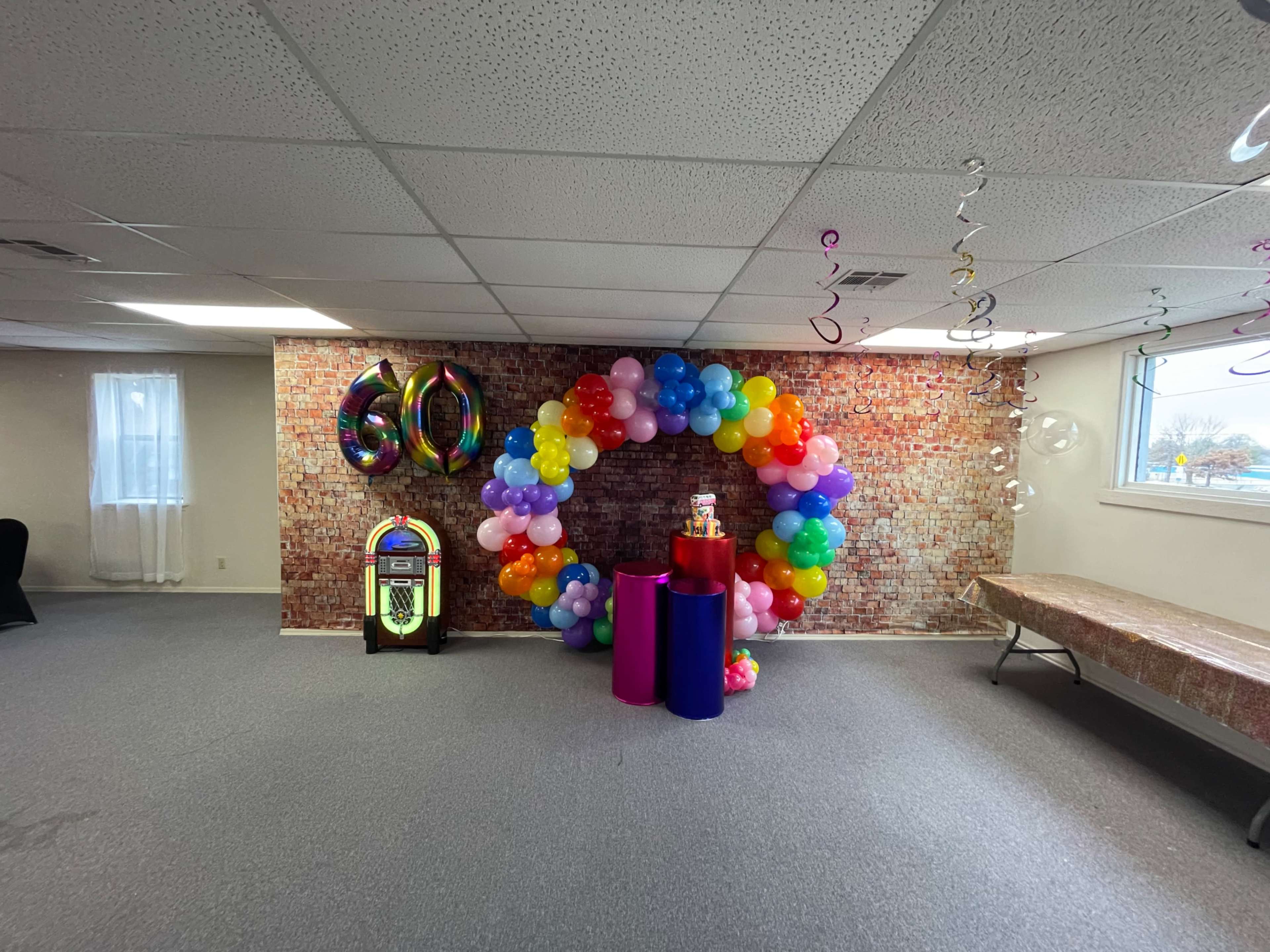 The image shows a decorated room with a balloon arch, a colorful display, a jukebox, and a large "60" balloon, indicating a celebration.