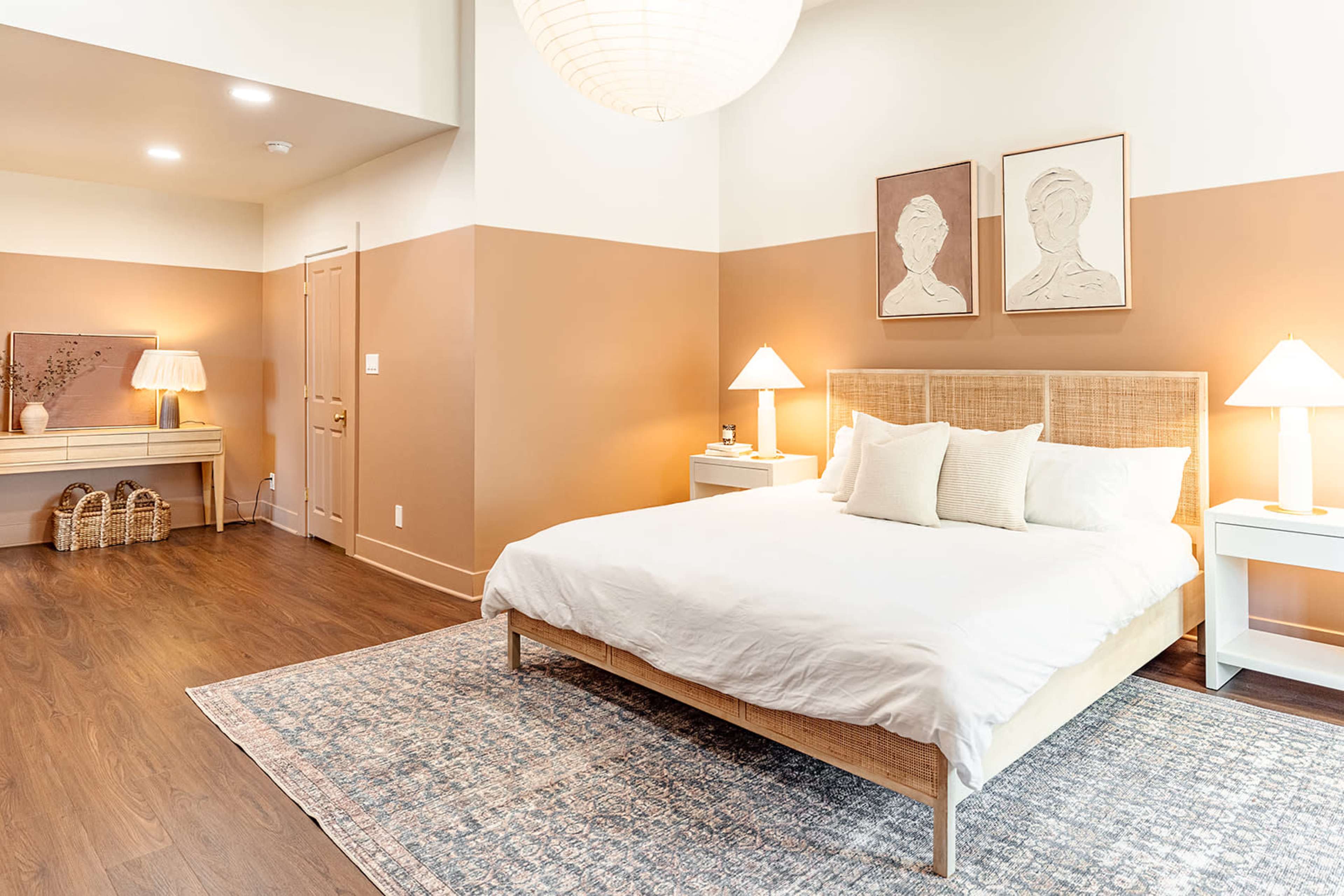 A well-lit bedroom features a large bed with white bedding, two bedside lamps, and framed artwork on the walls.