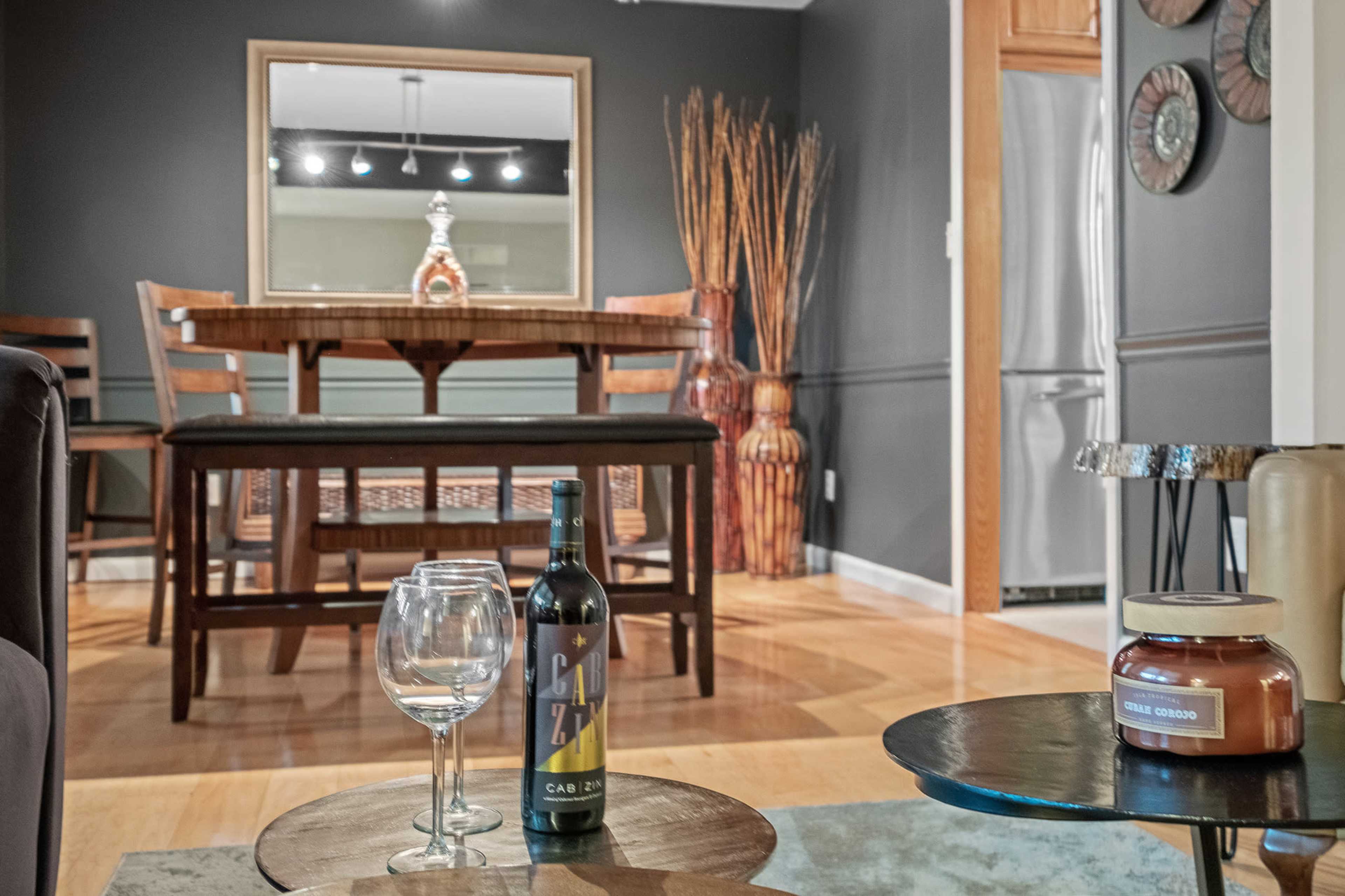 A modern dining area featuring a wooden table with chairs, a wine bottle and glasses on a foreground table, and decorative elements like a vase and an ornate mirror on the wall.