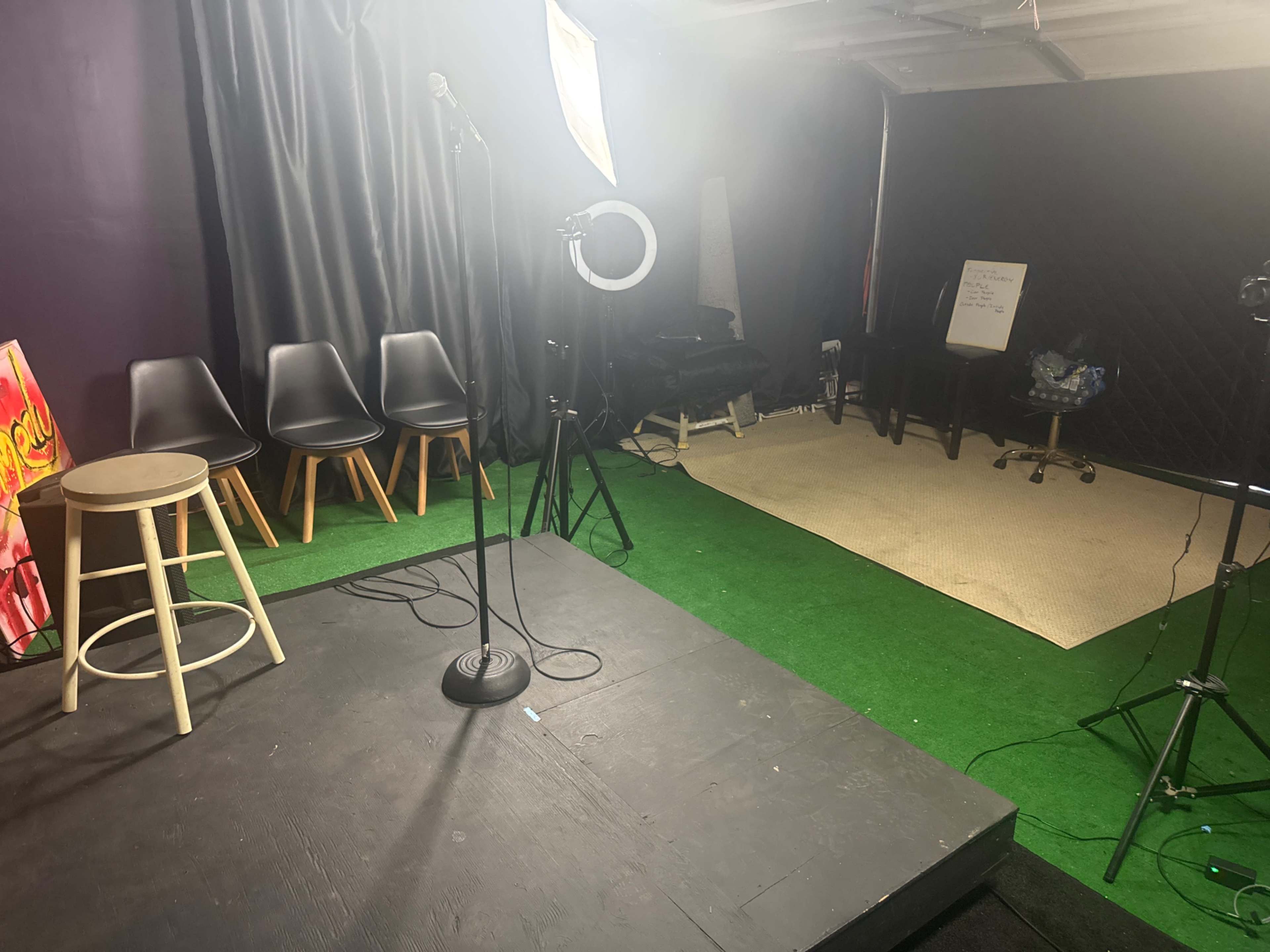 The image shows a small performance stage with a microphone stand, a stool, several chairs, and various lighting equipment in a room with black curtains and a green carpet.