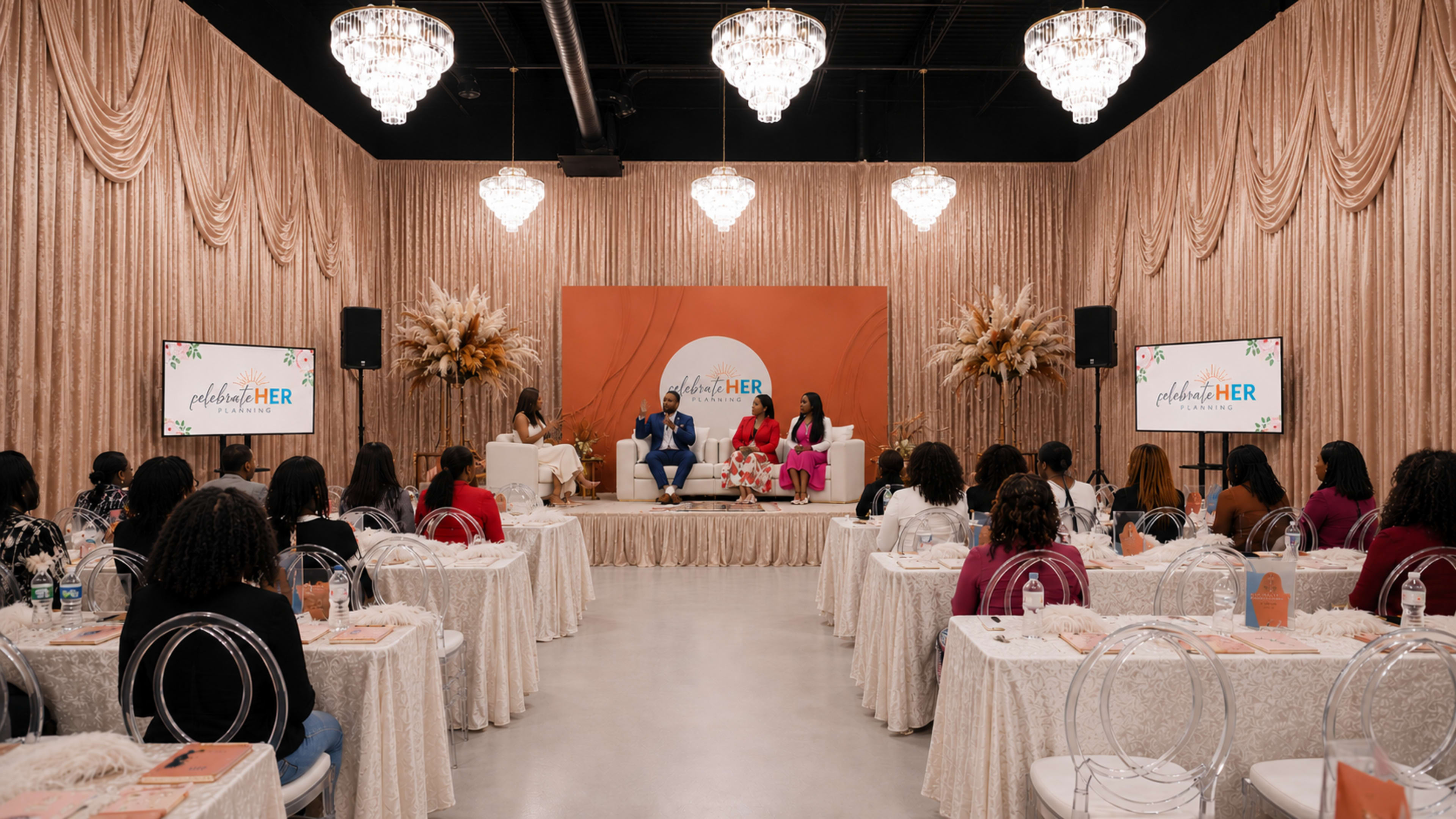 A panel discussion takes place on a stage in a well-decorated room, with an audience seated at tables, all under elegant chandeliers.