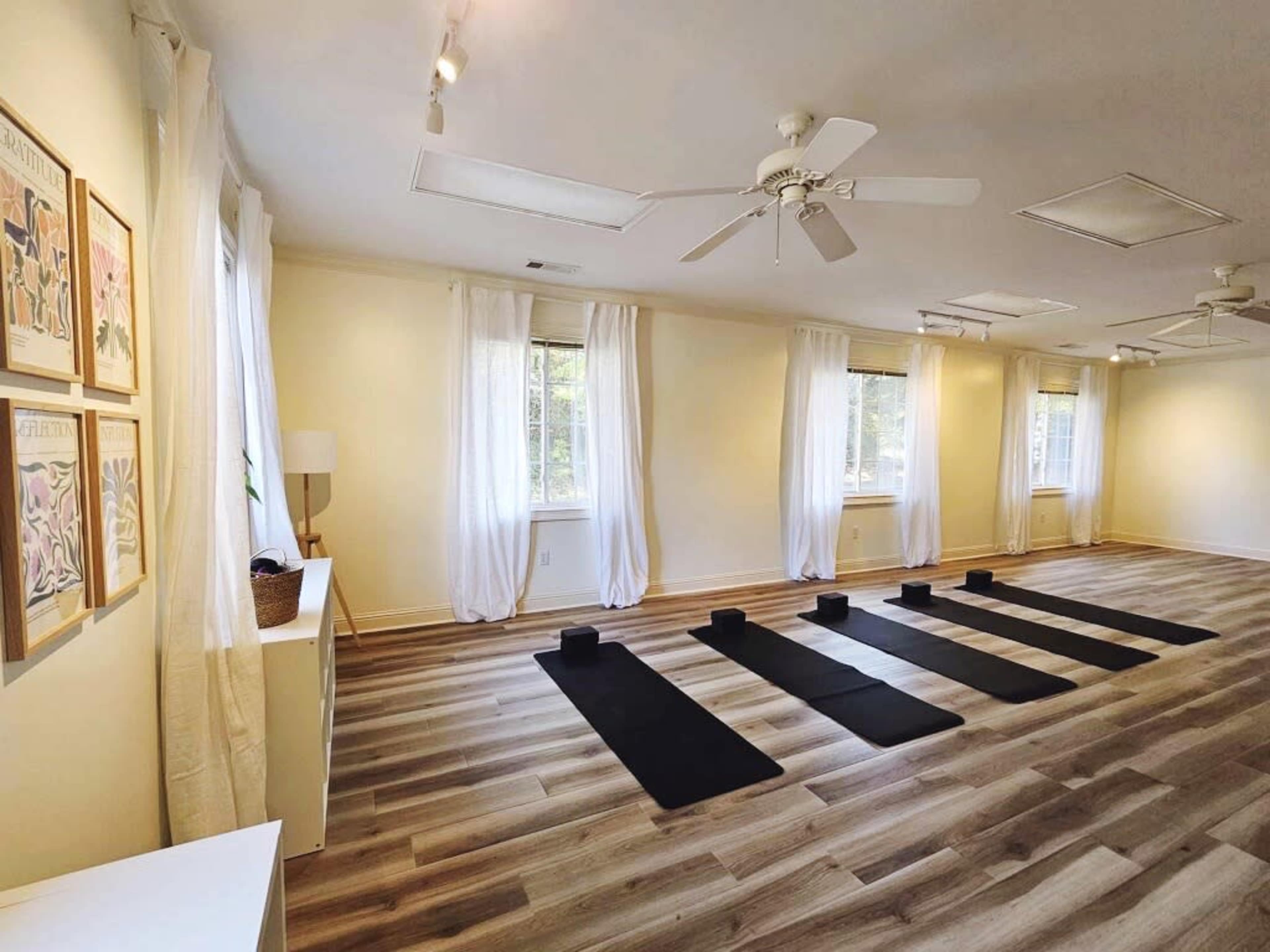 The image shows a bright, empty yoga studio with four yoga mats and blocks arranged in a row on a wooden floor.
