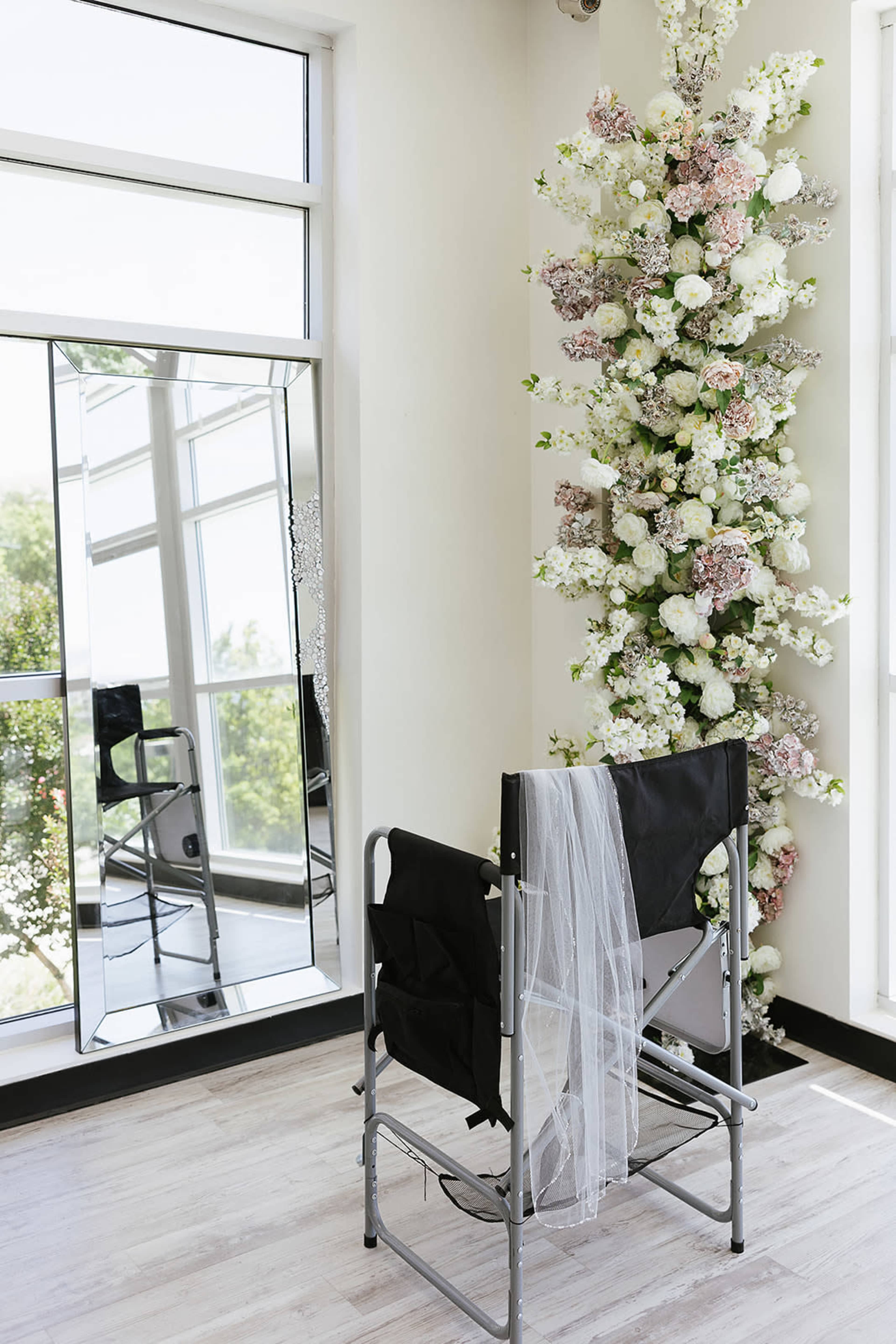The image shows a minimalist room with a black folding chair positioned in front of a large mirror, and a vertical arrangement of white and pastel flowers against the wall.